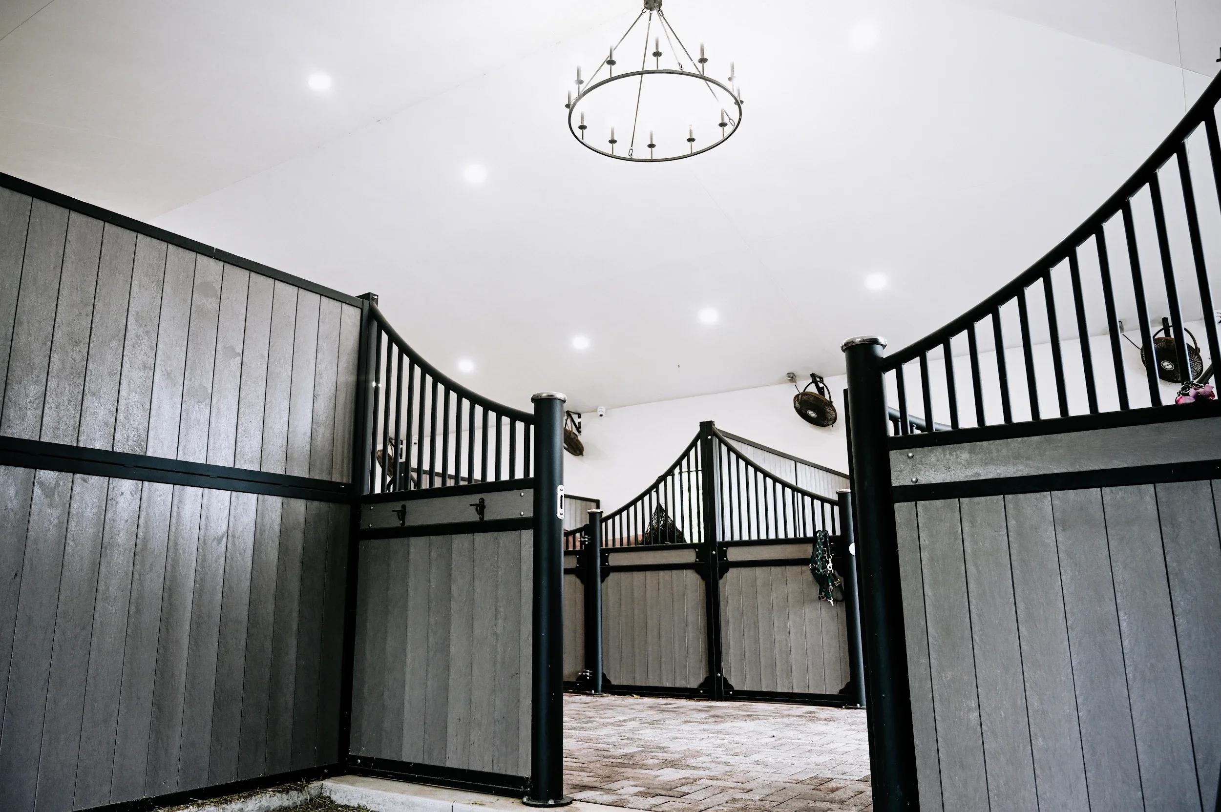 Interior of a riding arena with gray walls, black fences, white ceiling, chandelier, ceiling fans, and lighting