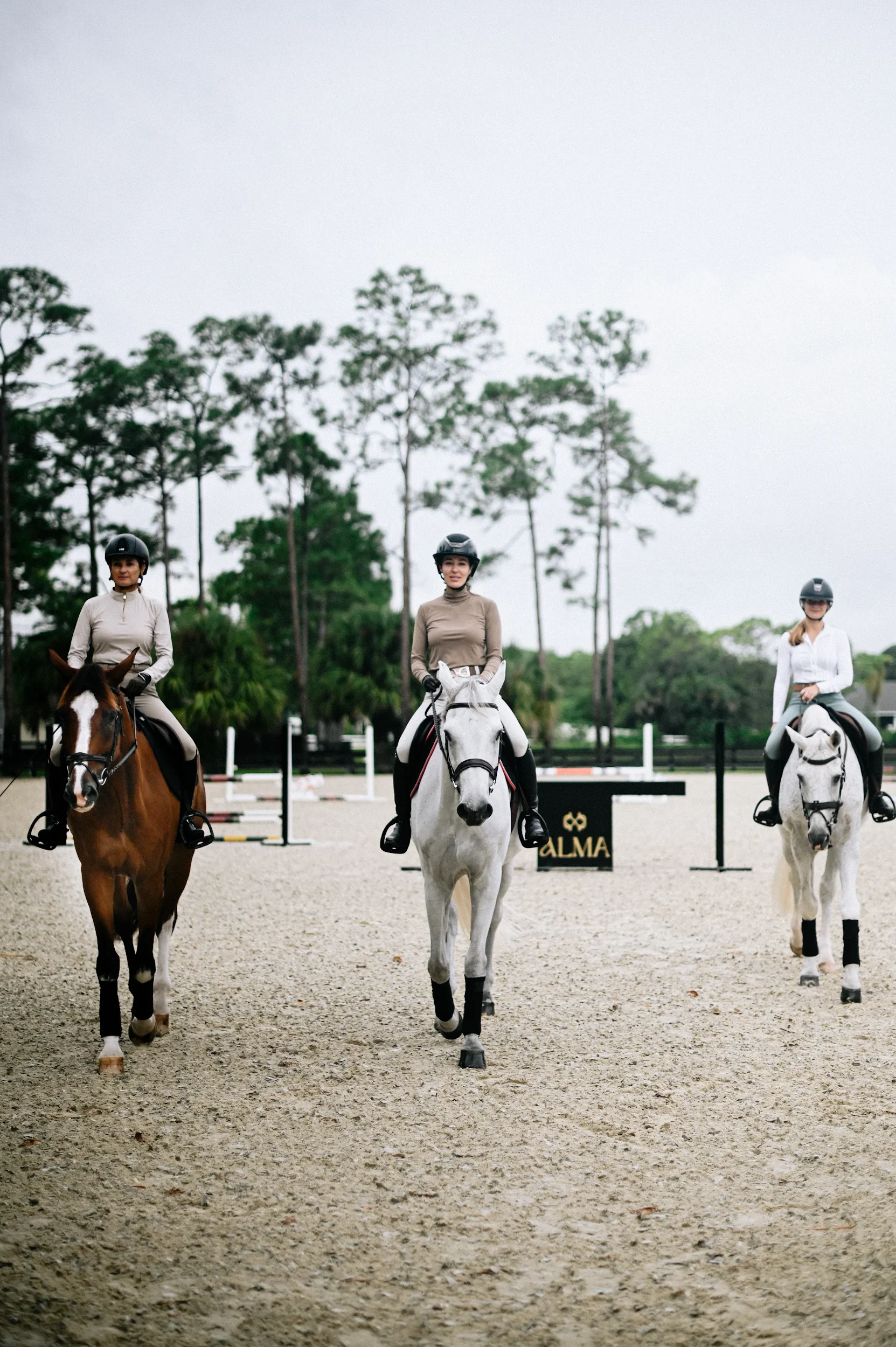 Three women riding horses in an outdoor equestrian arena, with jumps and trees in the background.