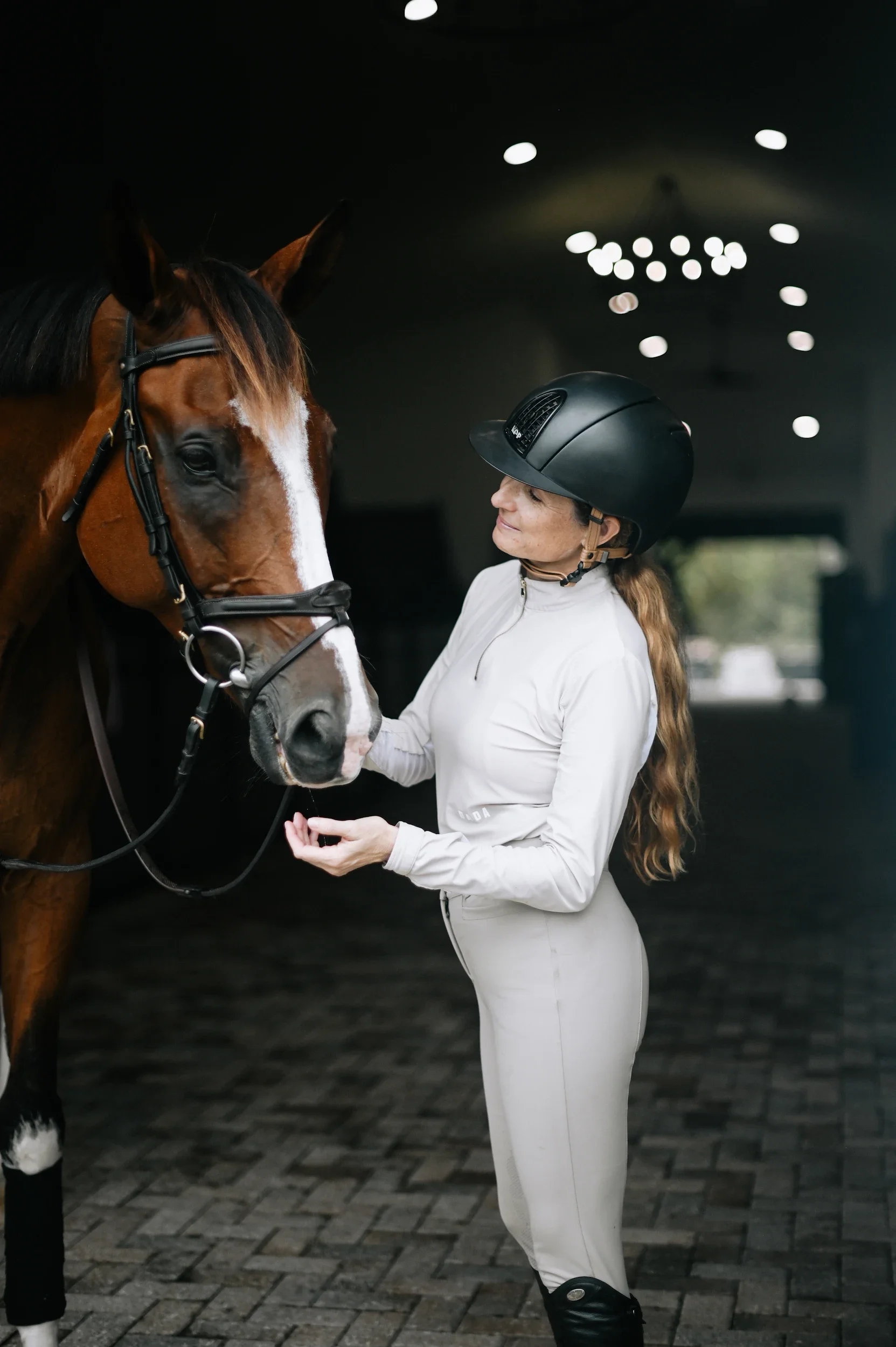 A woman in riding gear and a helmet softly touches a brown horse's face inside an indoor riding arena.