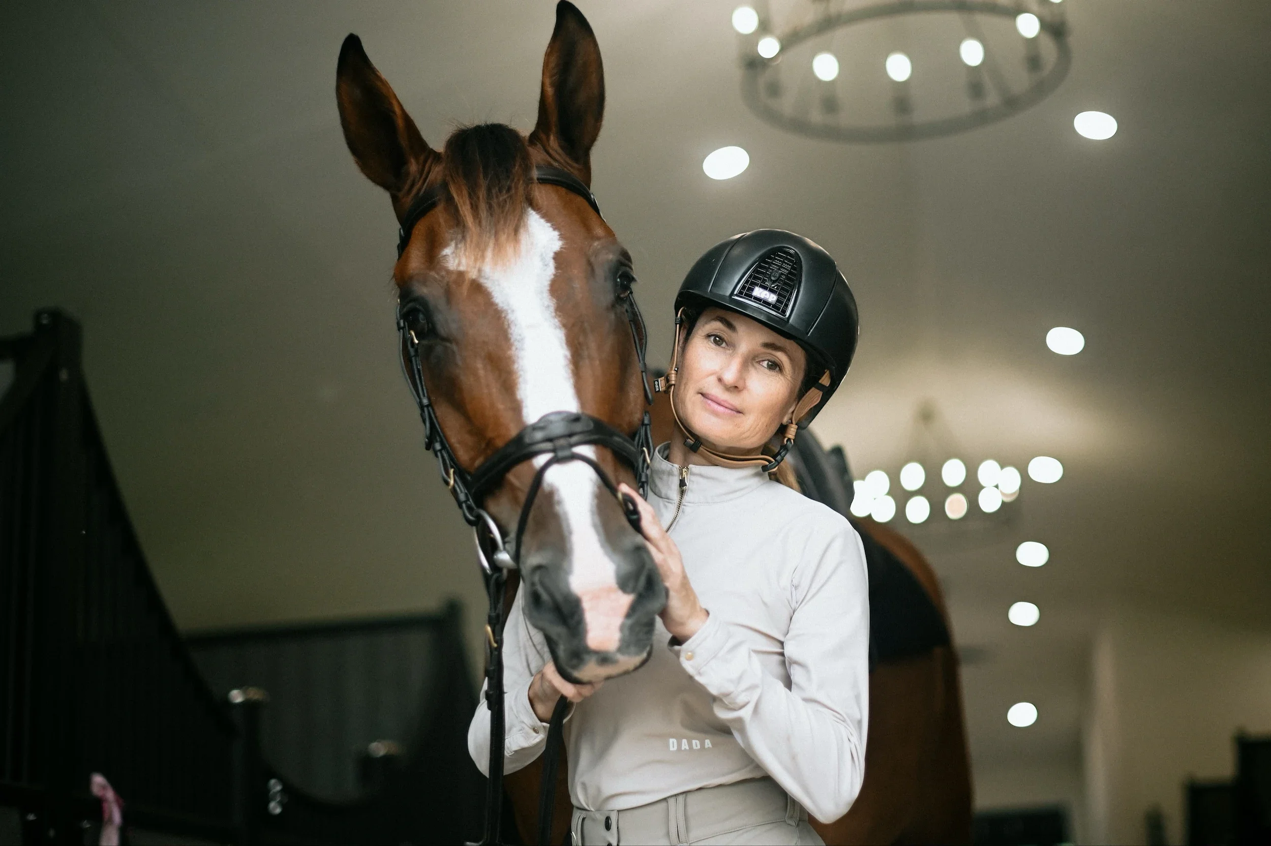 A woman in riding gear holding a brown horse with a white stripe on its face inside a stable or indoor riding arena.