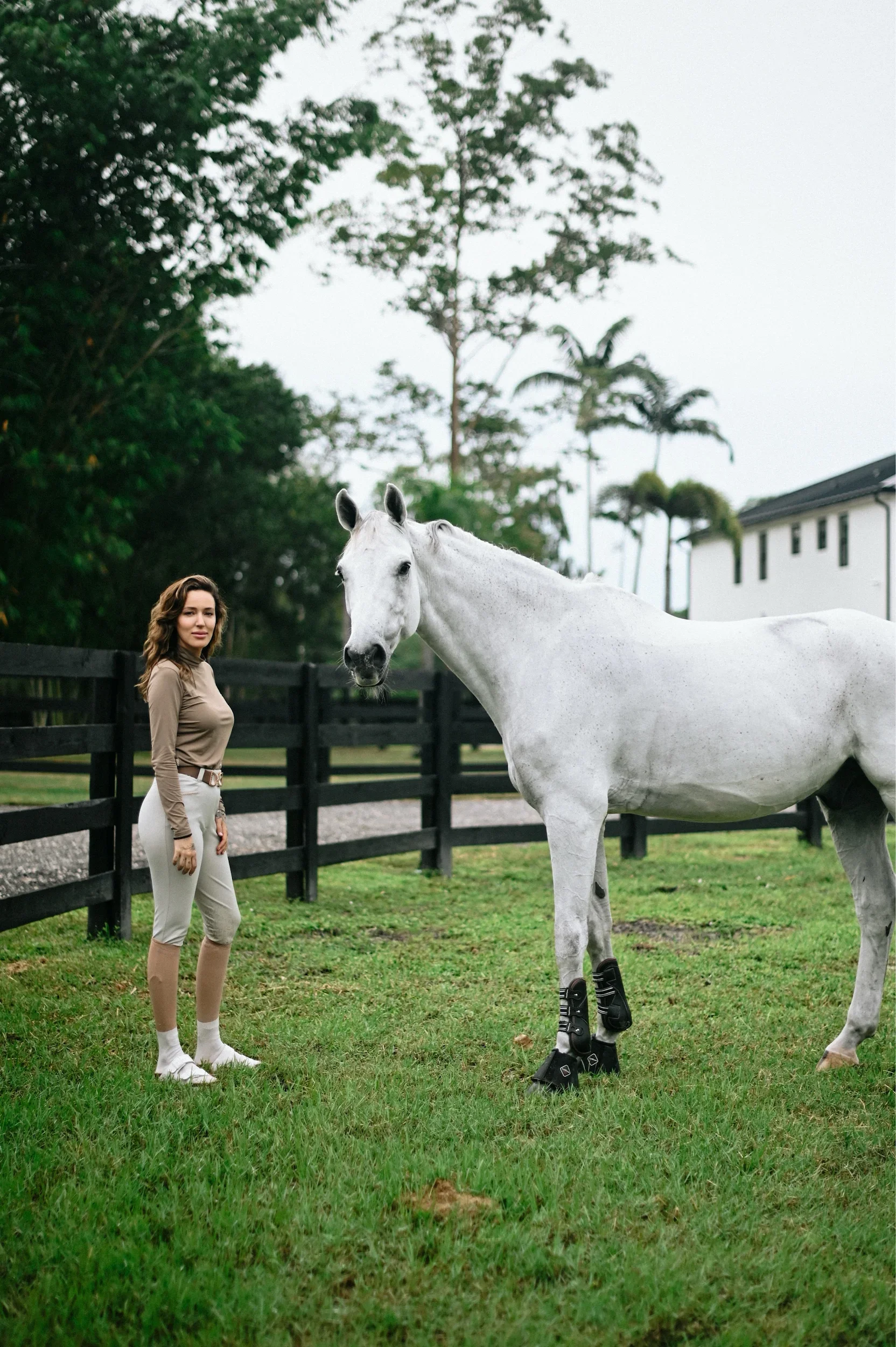 A woman standing next to a white horse in a grassy paddock with a black fence, trees, and a white building in the background.