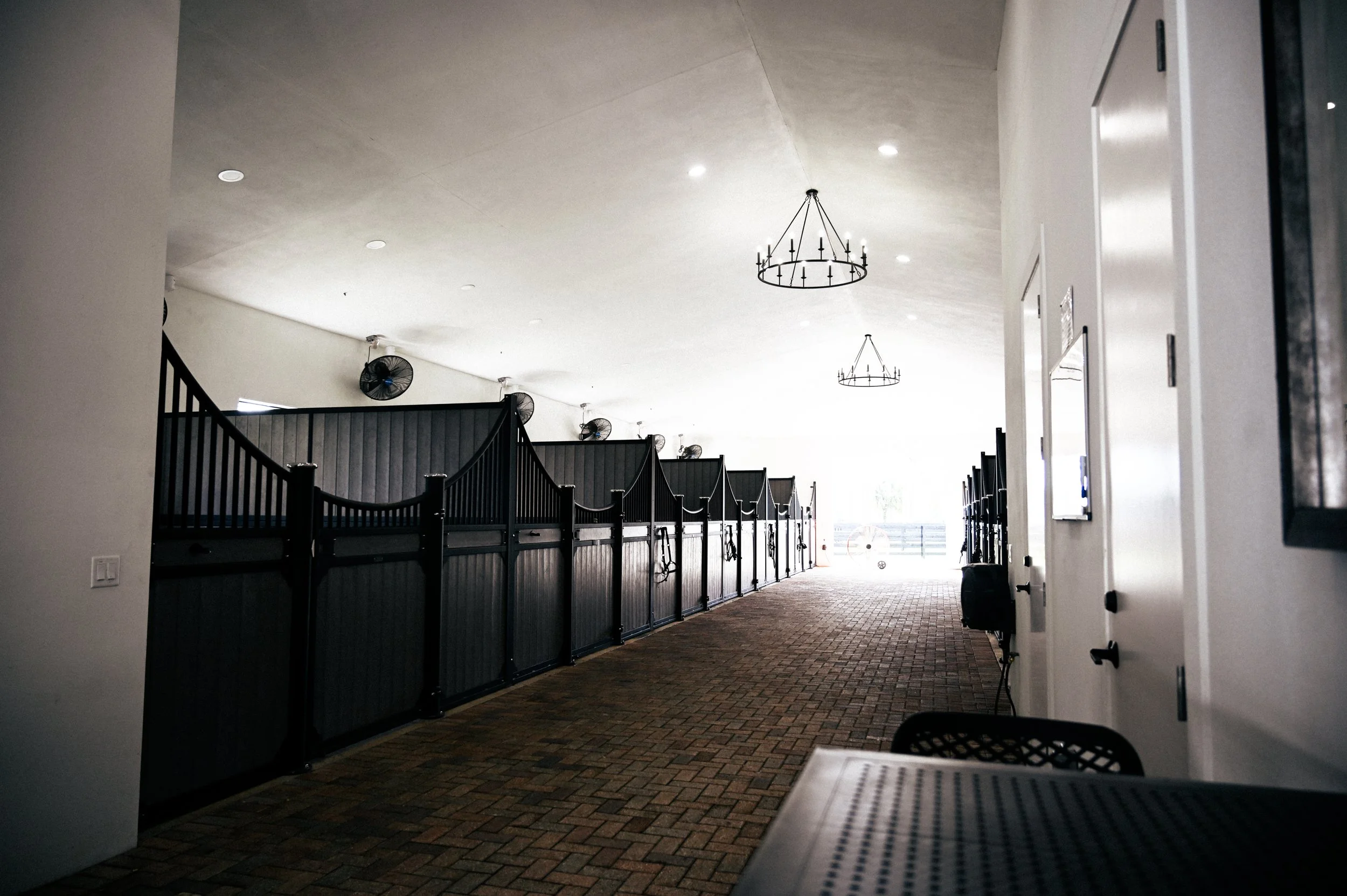 Indoor space with a brick floor, white walls, black barn-style stalls on the left, and white doors with black handles on the right. Several ceiling fans and chandeliers hang from the ceiling, with sunlight coming from an open area at the end of the r