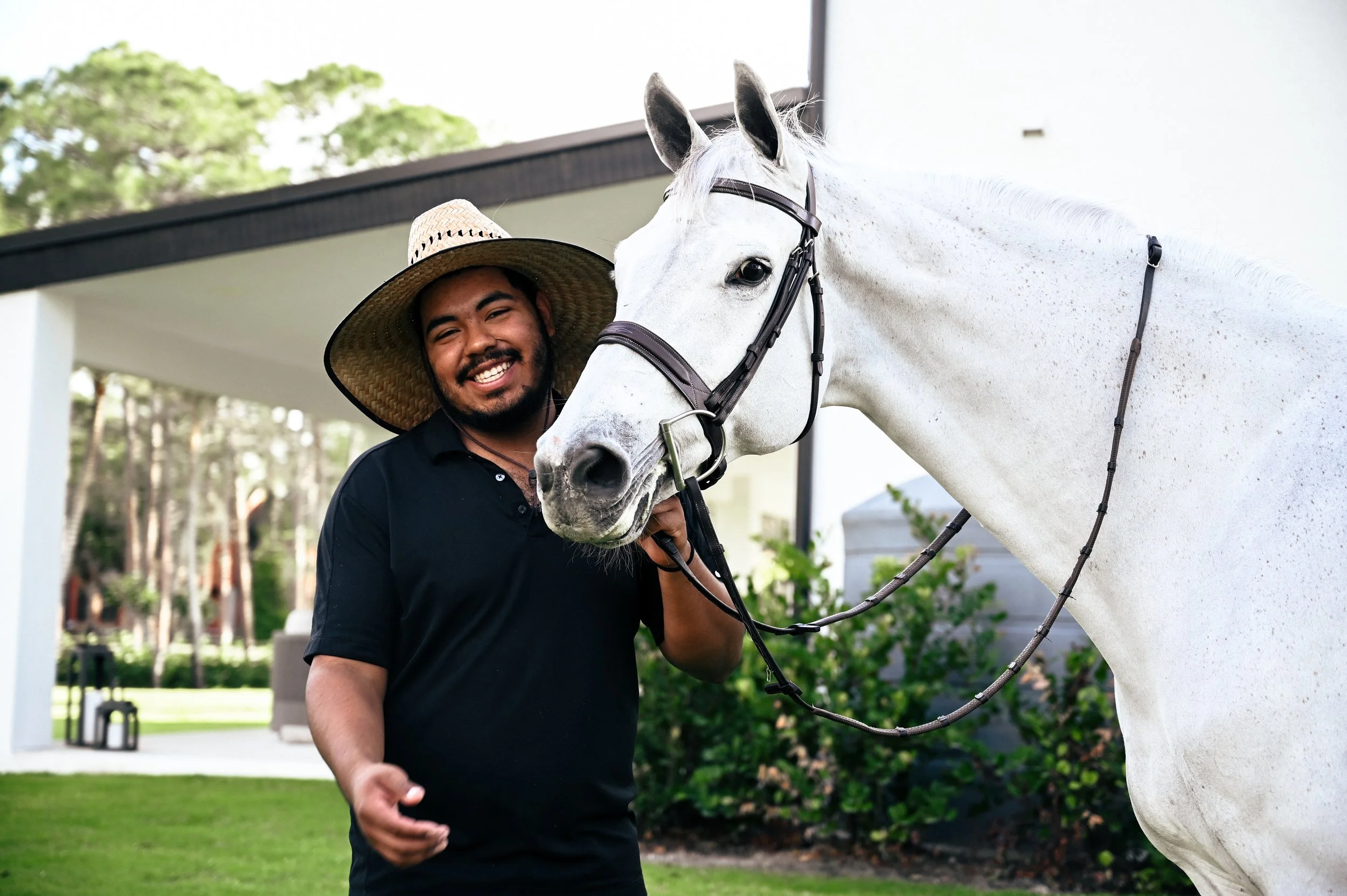 Smiling man with wide-brimmed straw hat standing next to a white horse outdoors.