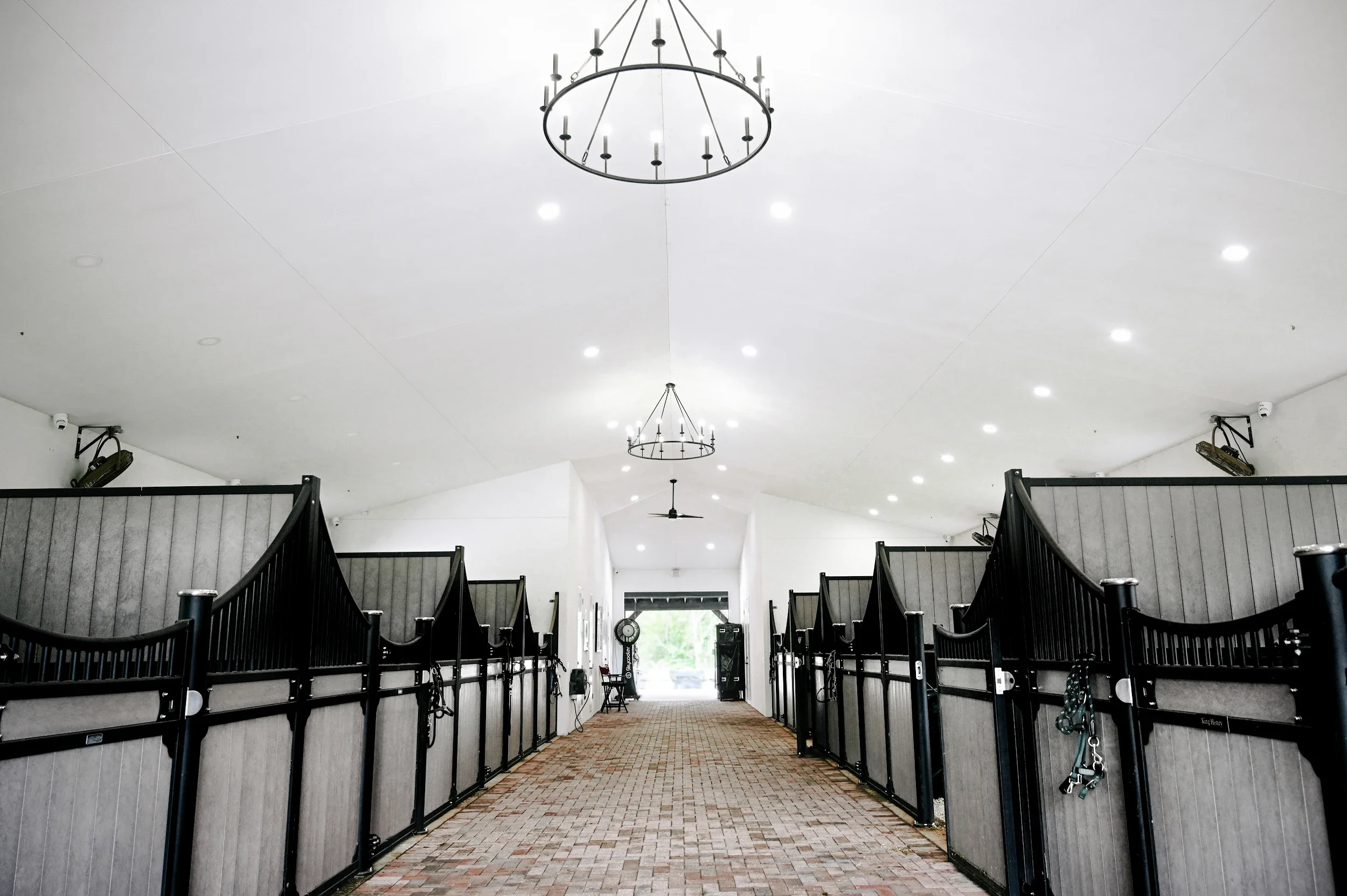 Inside view of a horse stable with black stall doors, a brick aisle, chandelier lights, ceiling fans, and a door with greenery outside.