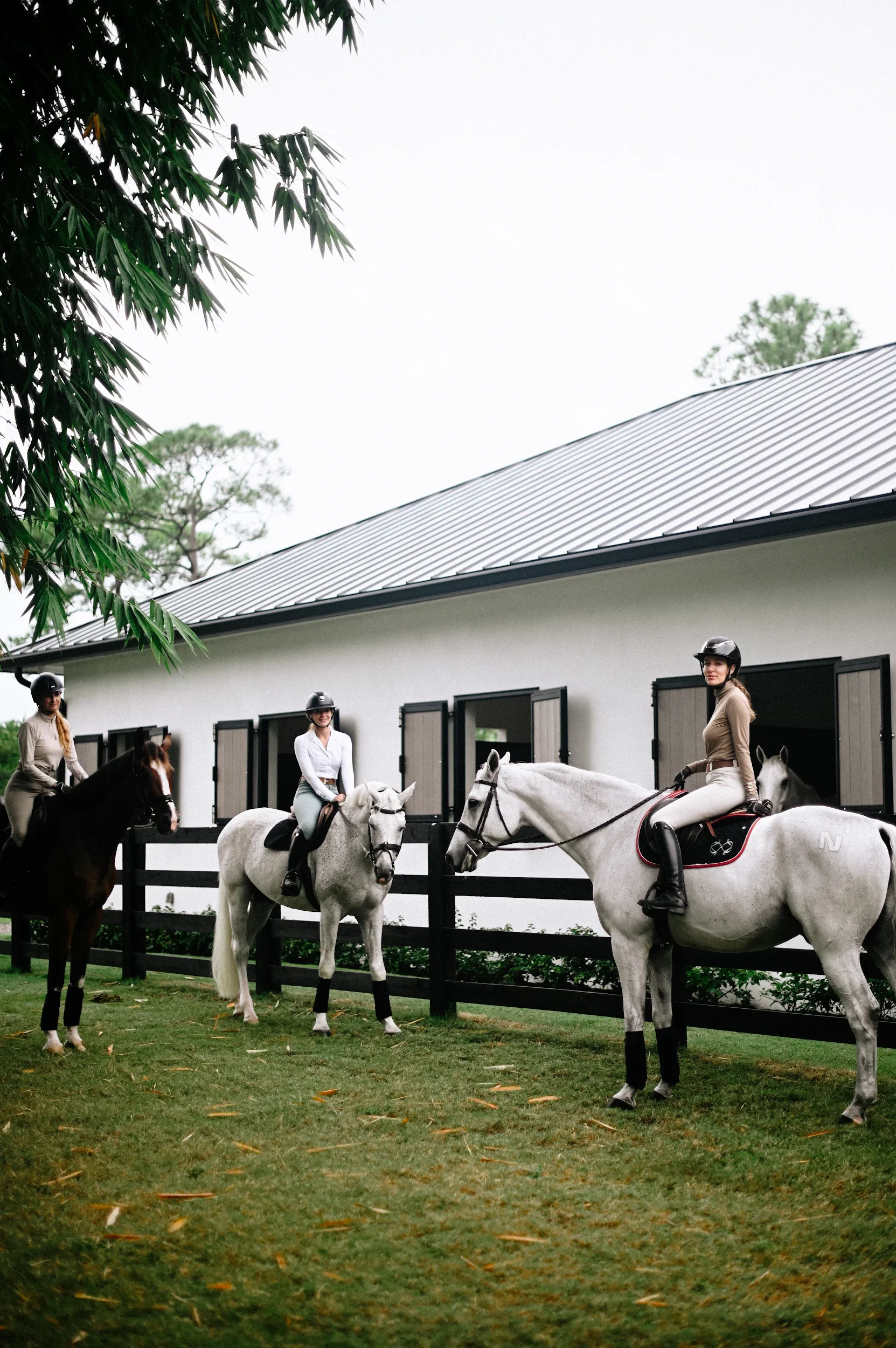 Three women riding horses along a black fence outside a white building with windows, with trees and a gray sky in the background.