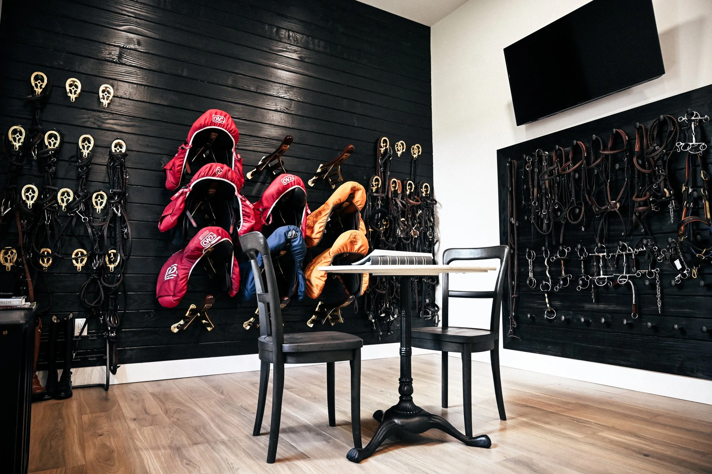 A room with black walls decorated with horse riding equipment, including bridles and saddles, and a small round table with two black chairs.