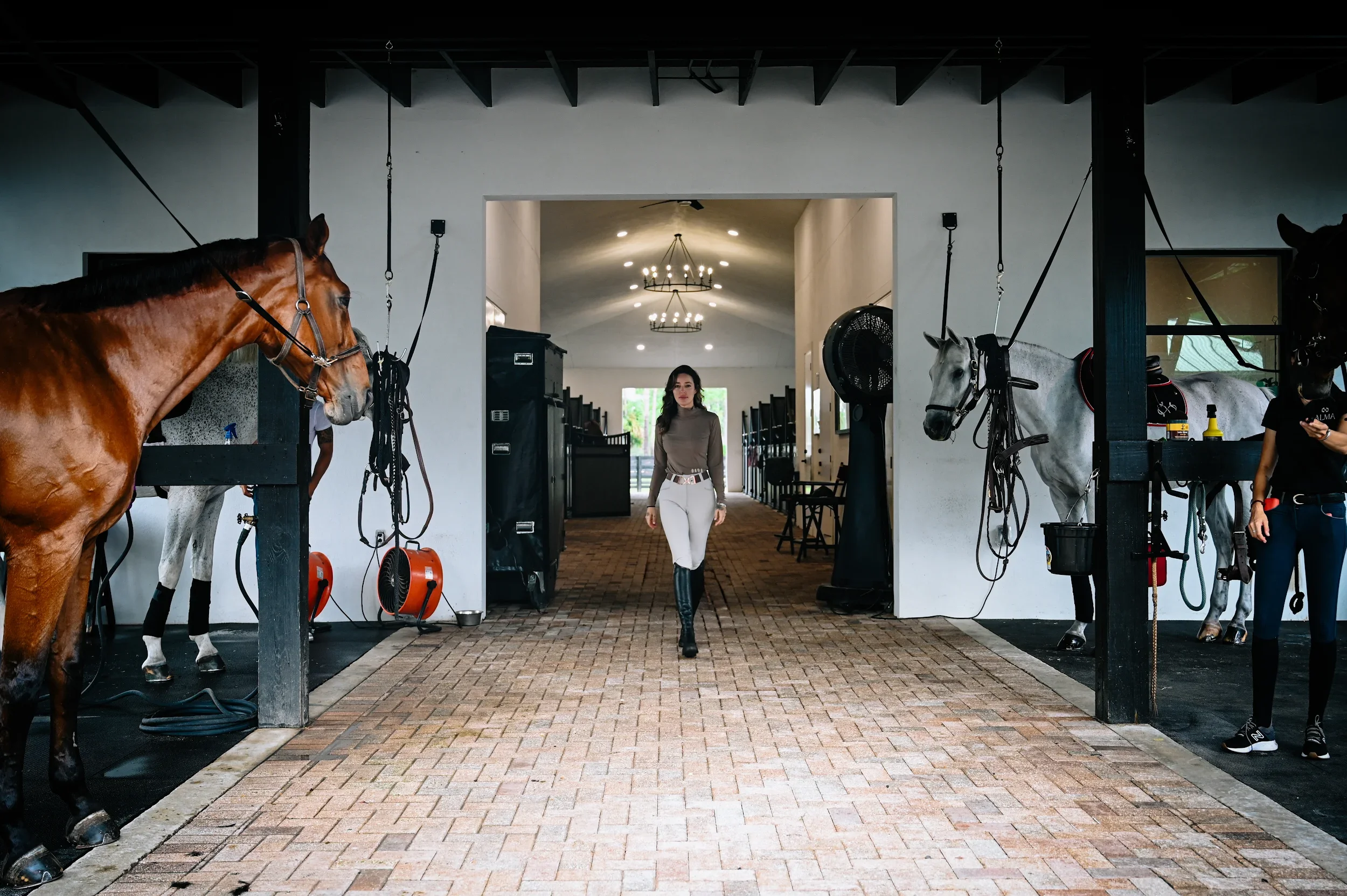 A woman walking through a stable aisle with horses in stalls on both sides.
