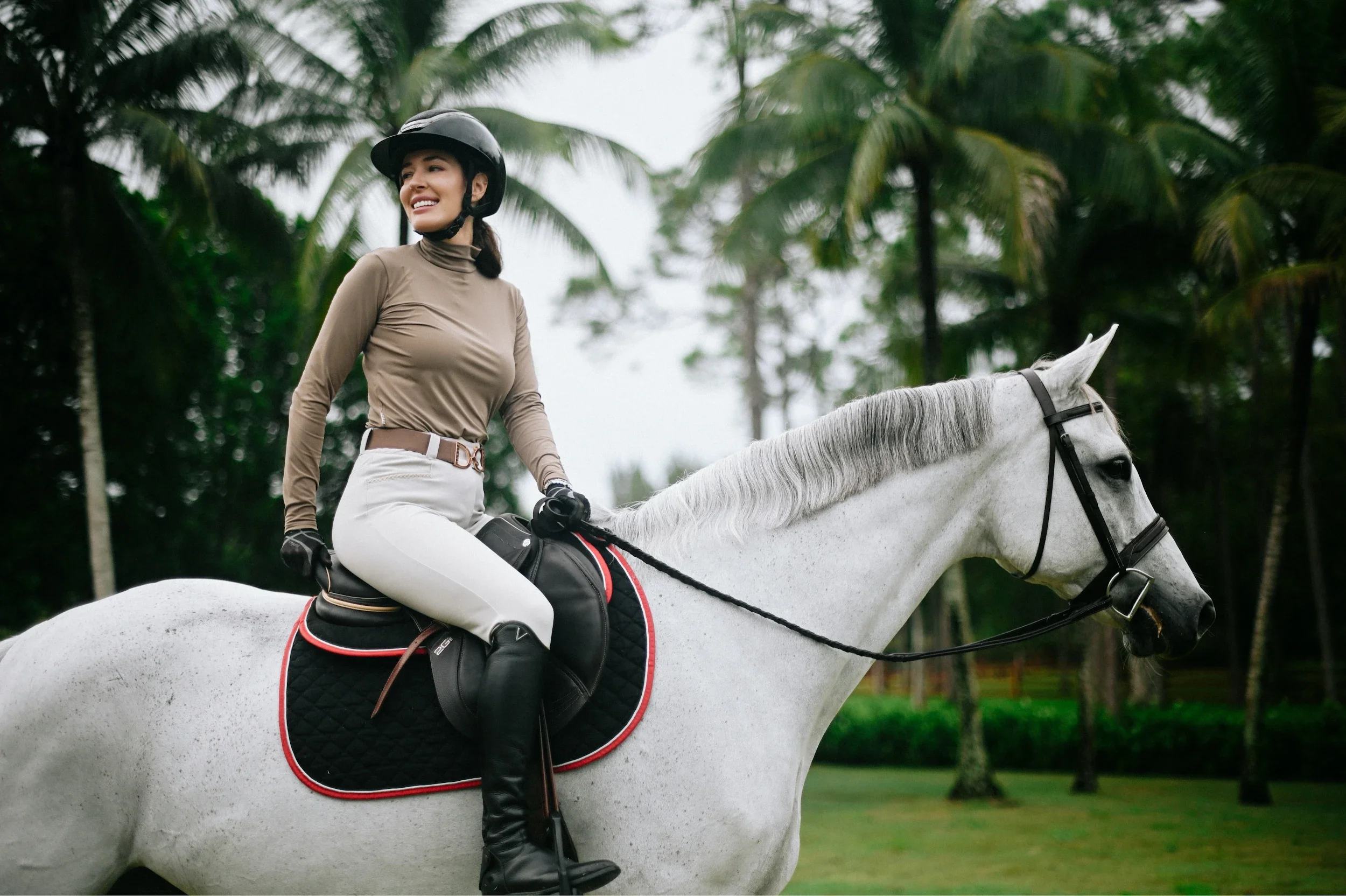 A woman in riding gear riding a white horse in a lush, green outdoor setting with palm trees