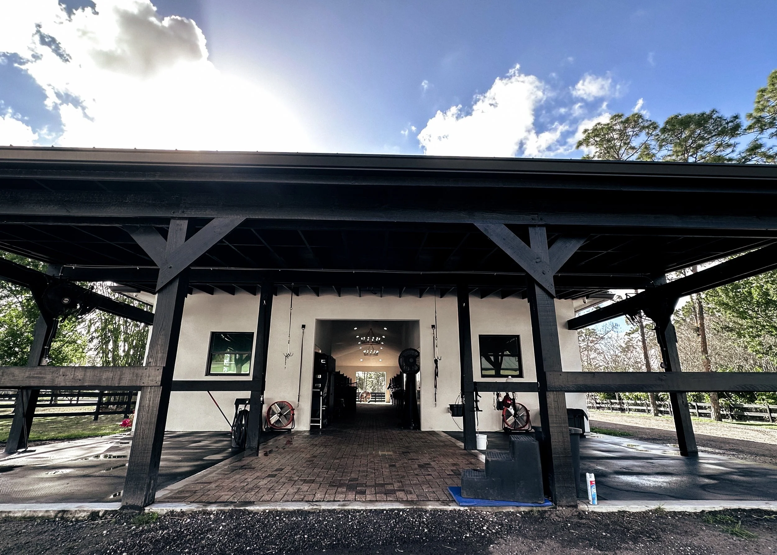 Front of a modern building with a large overhanging roof, black wooden beams, and an open entrance showing a glimpse of the interior, surrounded by trees and a clear blue sky.