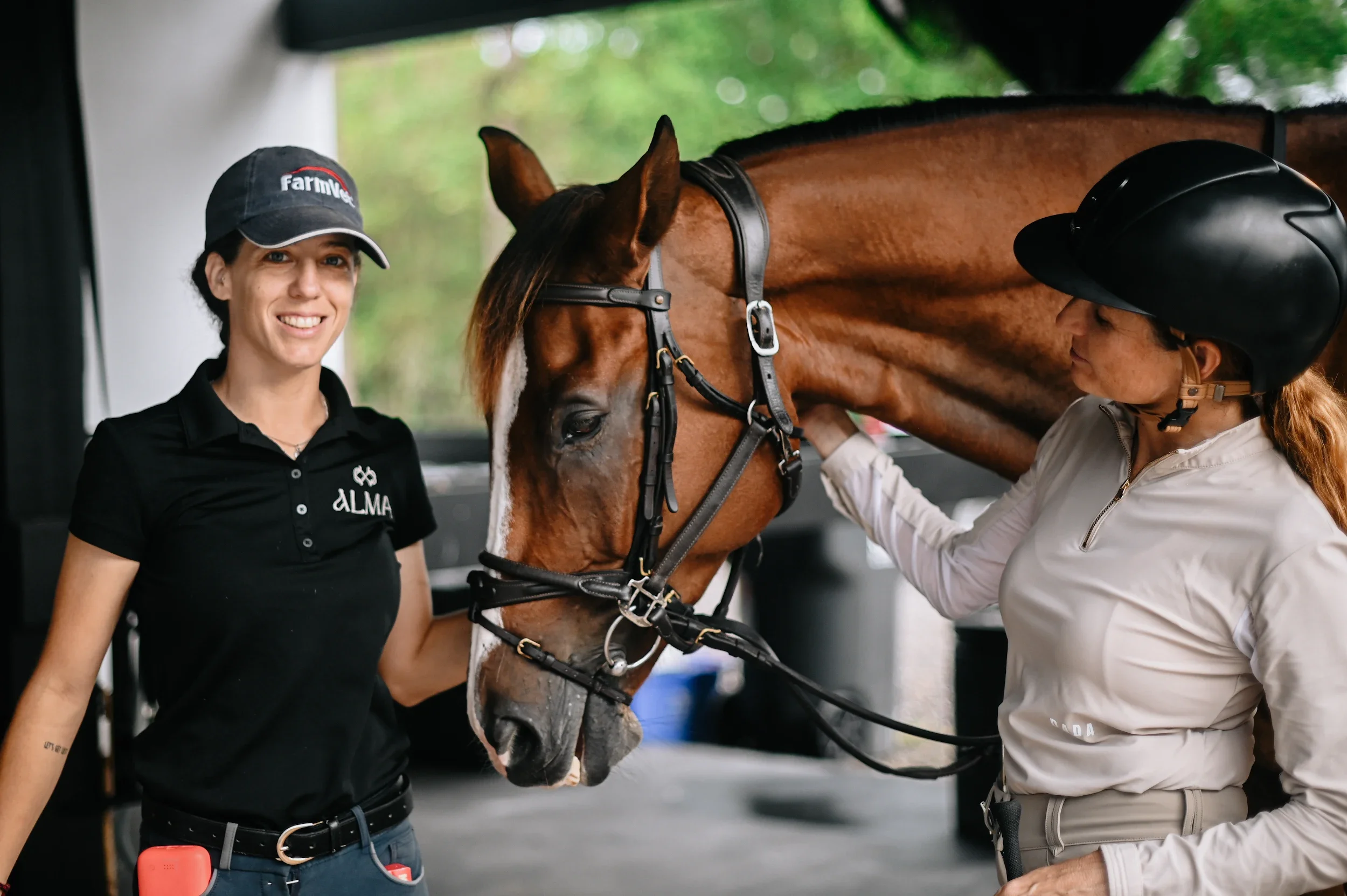 Two women and a horse inside a stable. One woman, in a black shirt and cap, is smiling and holding the horse's bridle. The other woman, wearing a helmet and riding gear, is gently touching the horse's neck, which has a shiny chestnut coat.