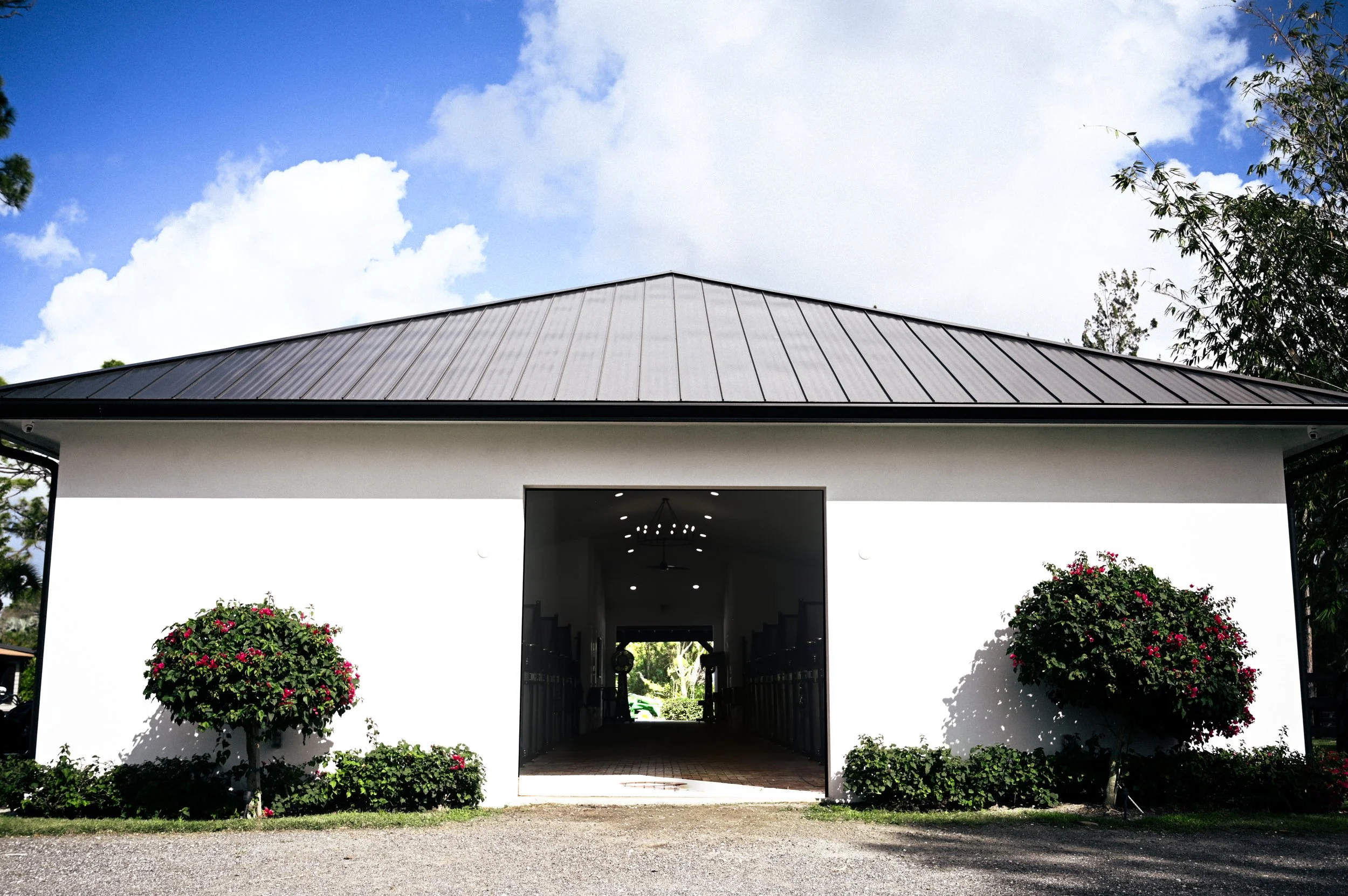 A white building with a dark metal roof, open entrance, symmetrical bushes with pink flowers on each side, and a gravel path leading to the entrance, under a partly cloudy sky.