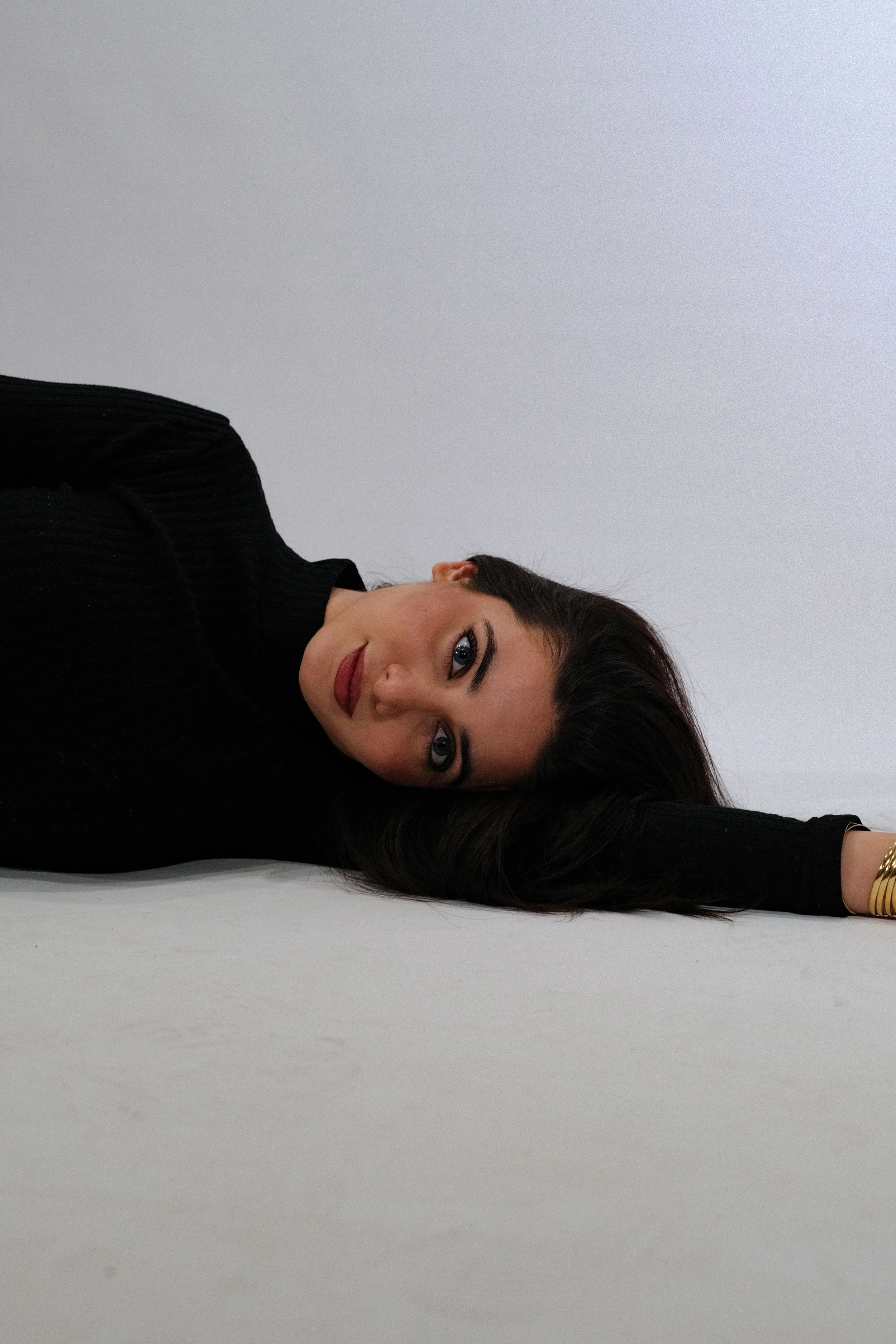 A woman with long dark hair and blue eyes lying on her side on a white surface, resting her head and arm on the ground, wearing a black long-sleeve top and gold bracelet.