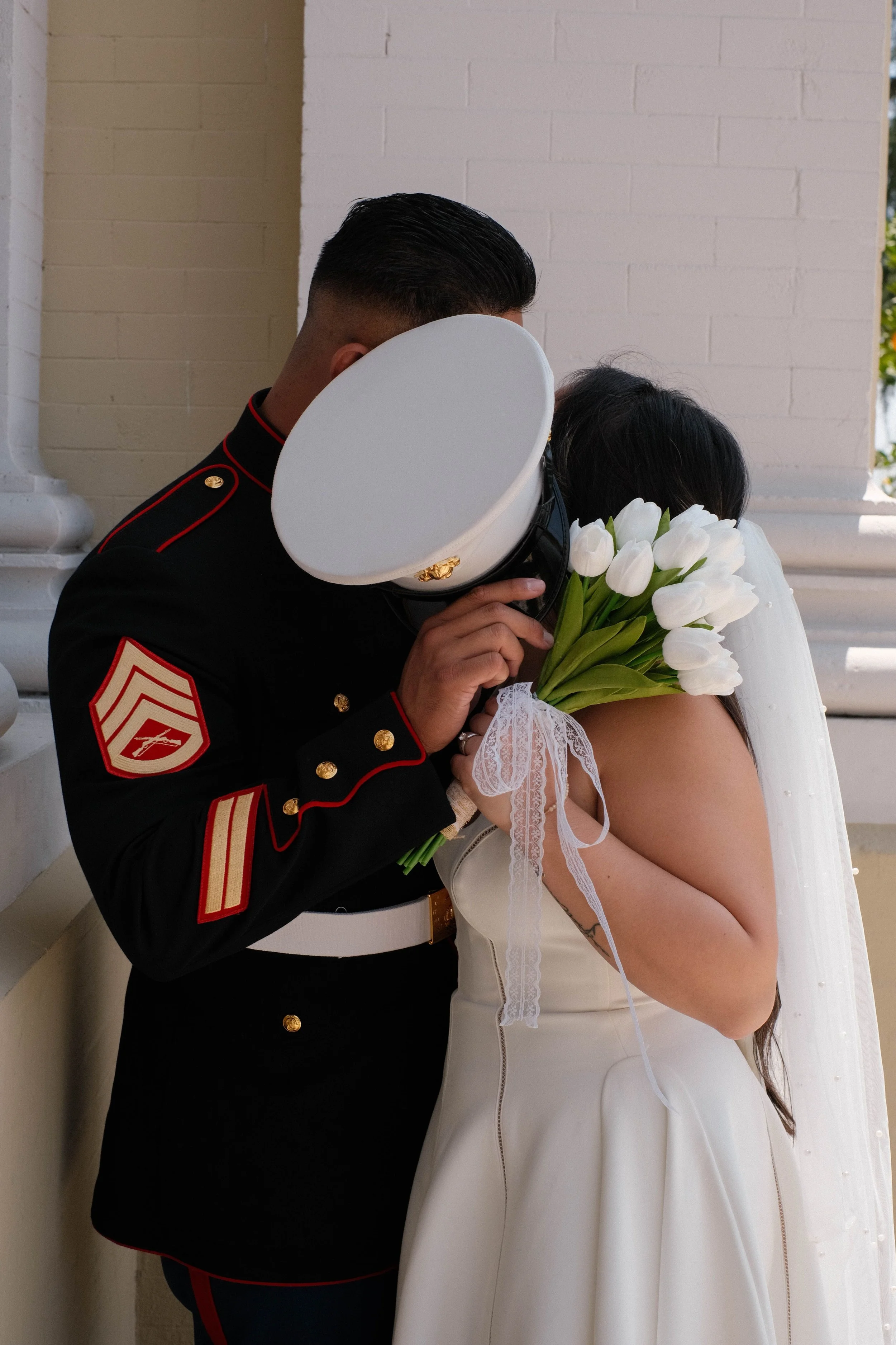 A U.S. Marine in dress uniform holding a bouquet of white tulips and a woman in a white wedding dress resting her head on his shoulder during a wedding moment.