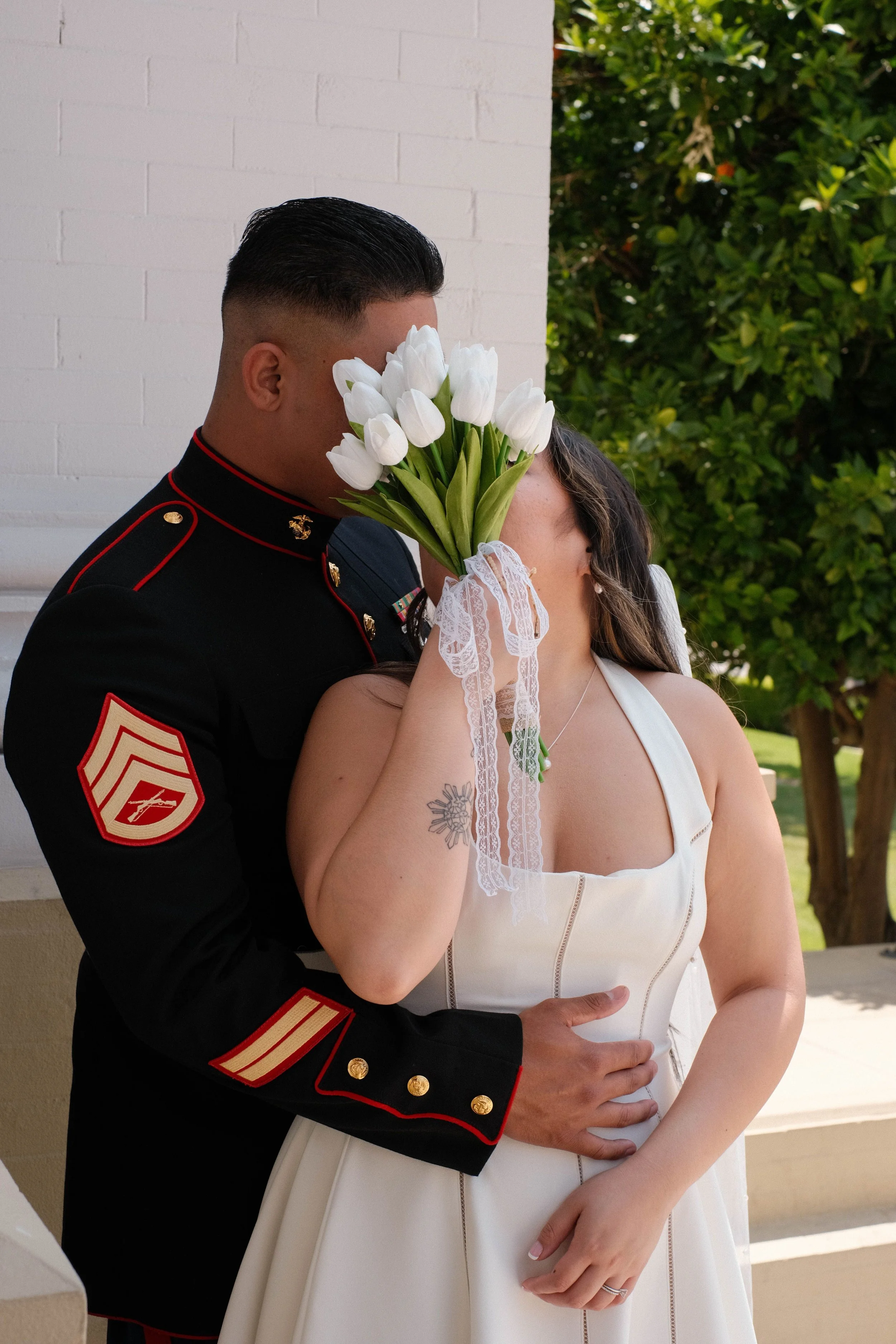A couple sharing a kiss with the woman holding a bouquet of white tulips in front of her face, outdoors on a porch with greenery in the background.
