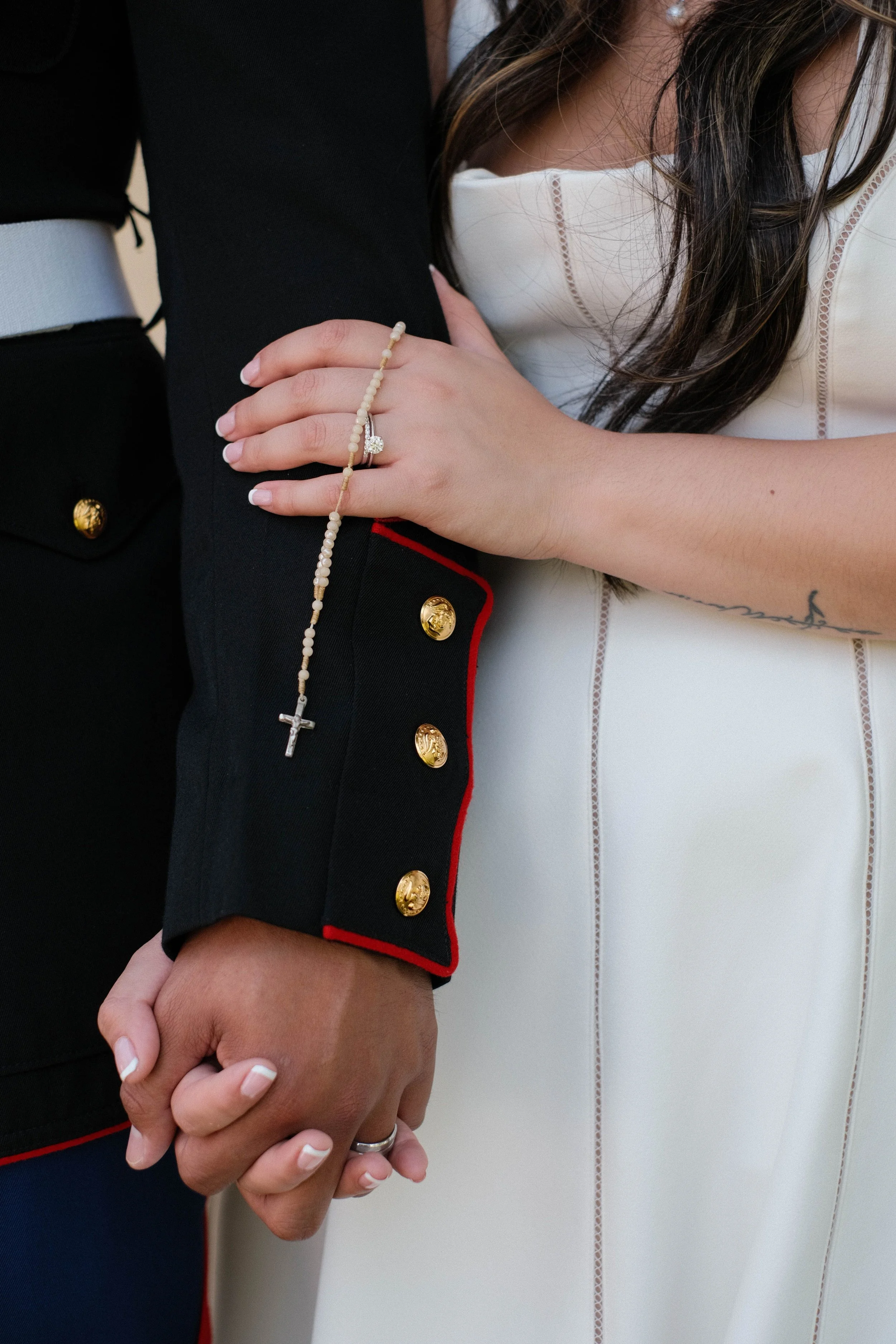 A couple holding hands, with one person wearing a black uniform with gold buttons and a white piping edge, the other wearing a white dress with visible stitching, and a rosary hanging from the hand.