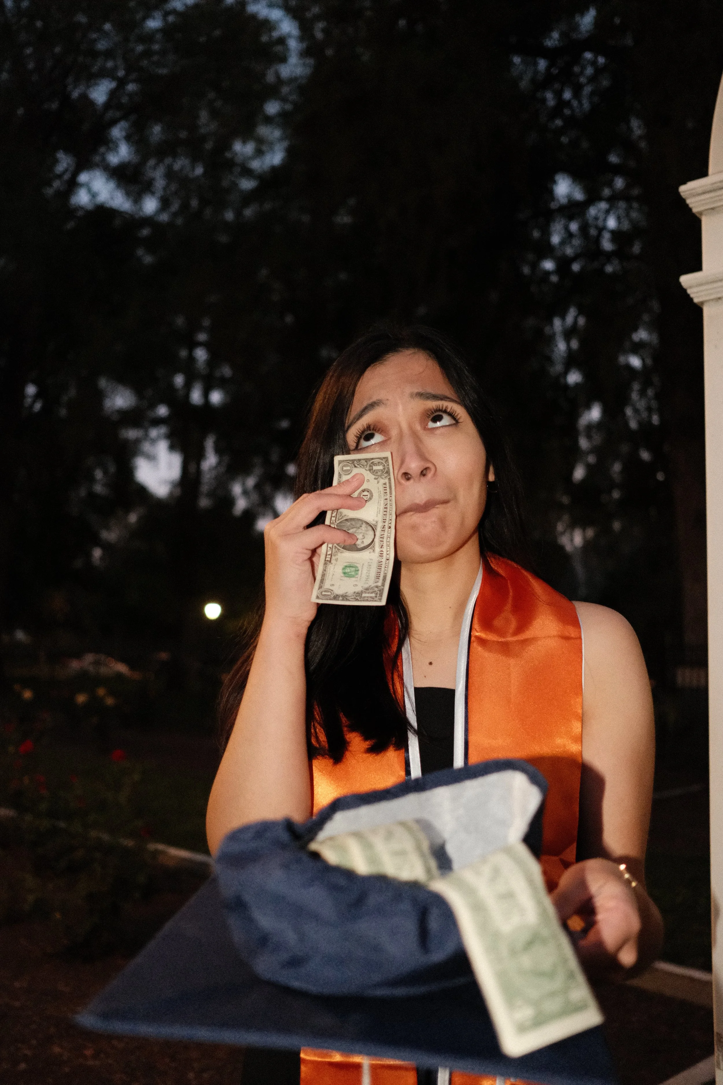 A woman in an orange graduation stole is holding money, with some bills in her hand and a few in a bag, standing outdoors during evening or night.