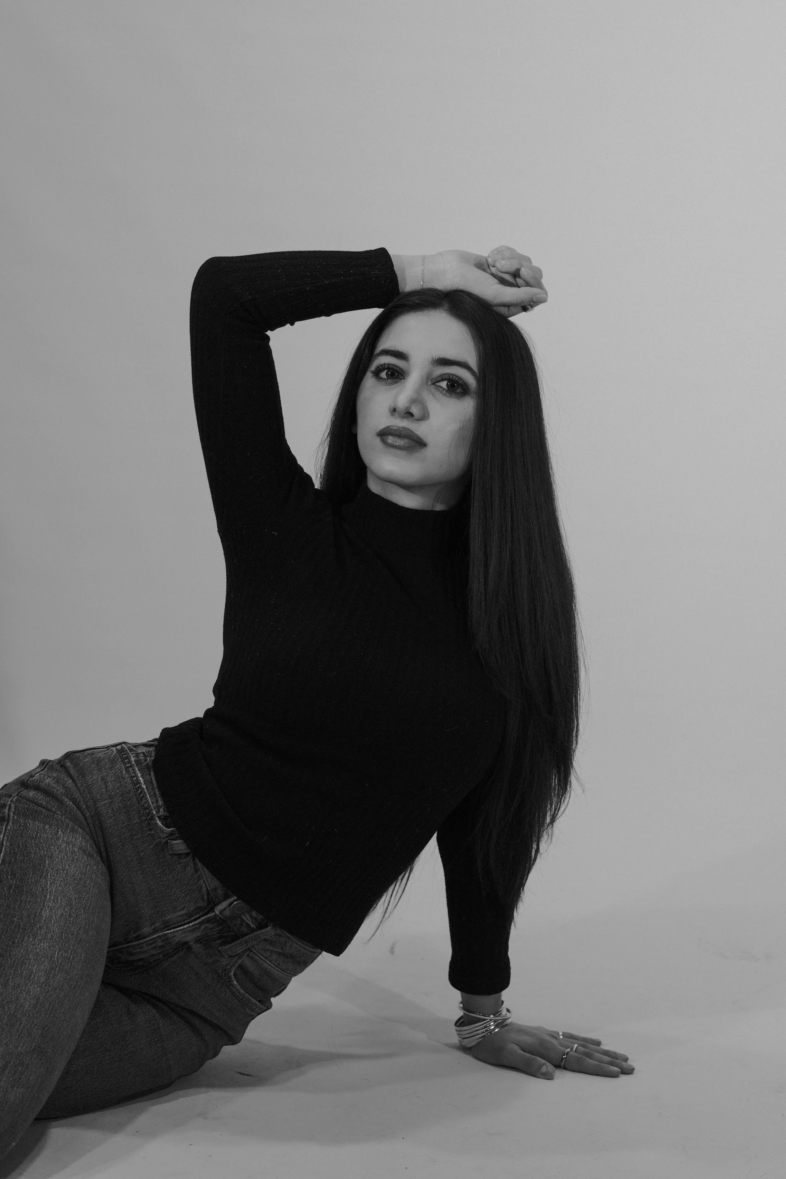 A woman with long dark hair, wearing a black turtleneck and jeans, posing against a plain wall. She is leaning on one arm with her other arm raised above her head, with a confident expression.