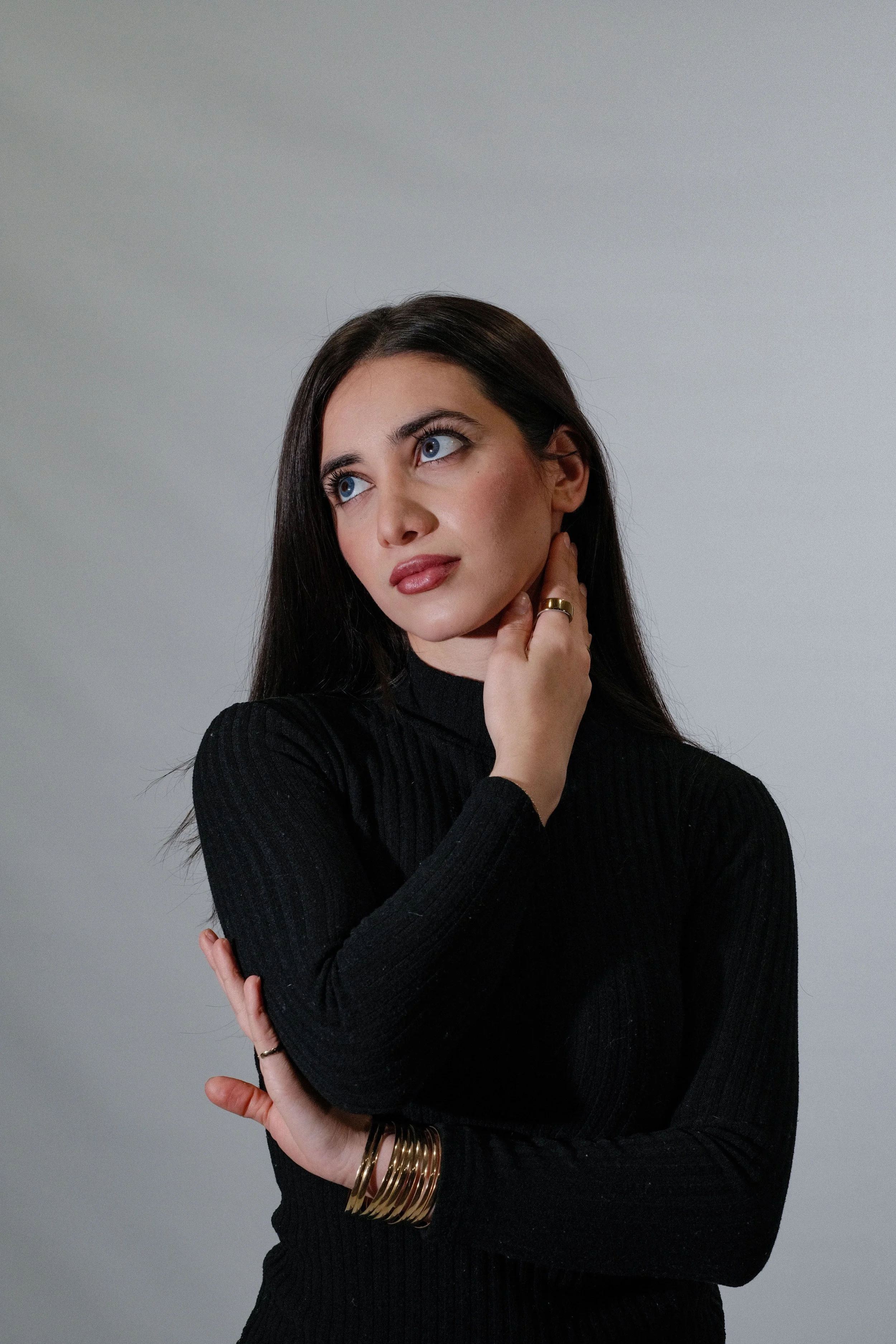 A woman with long dark hair and blue eyes looking thoughtfully to the side, wearing a black long-sleeve top and multiple gold bangles on her left wrist, posing against a plain gray background.