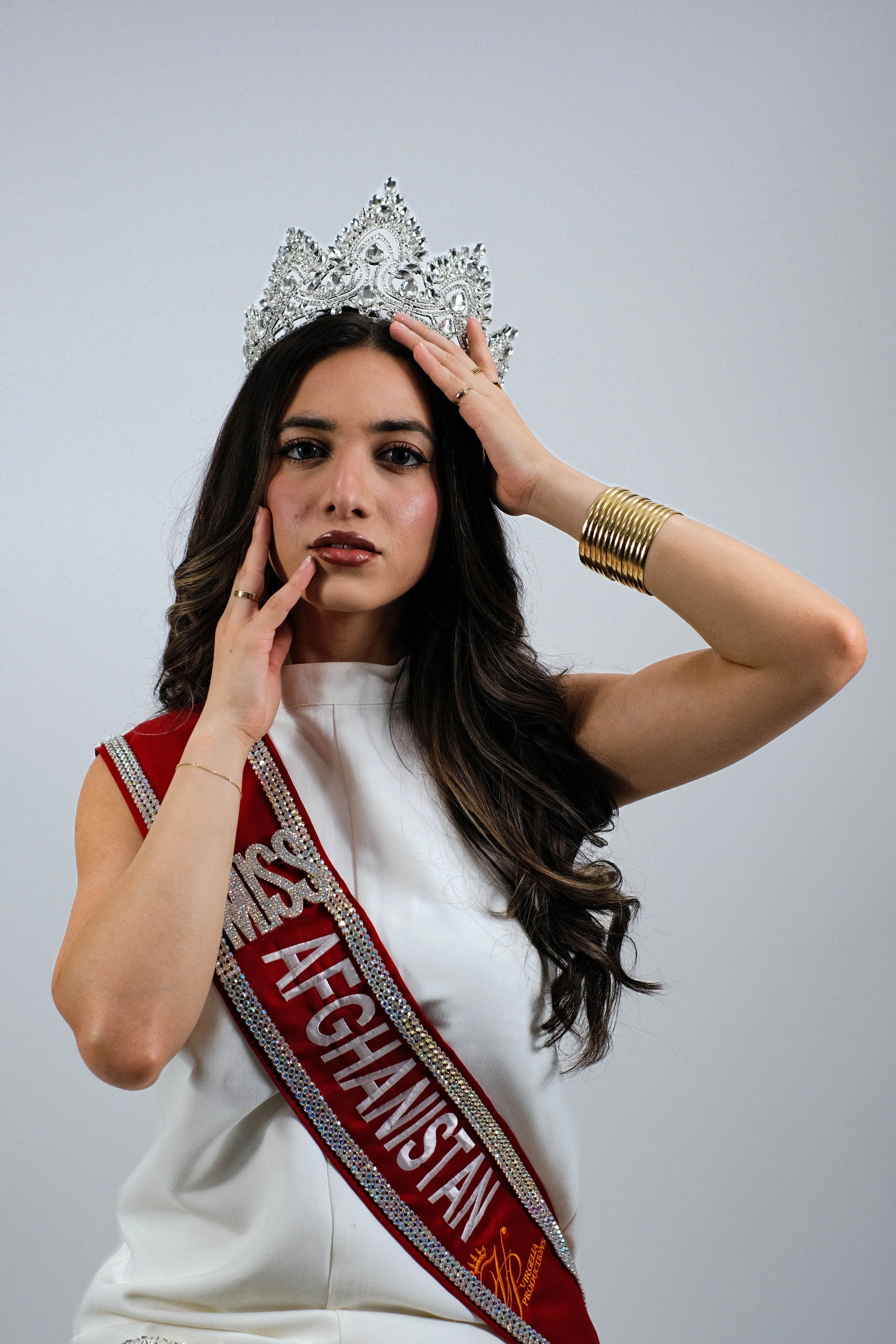 Miss Afghanistan wearing a crown, sash, and gold jewelry, standing against a plain background.