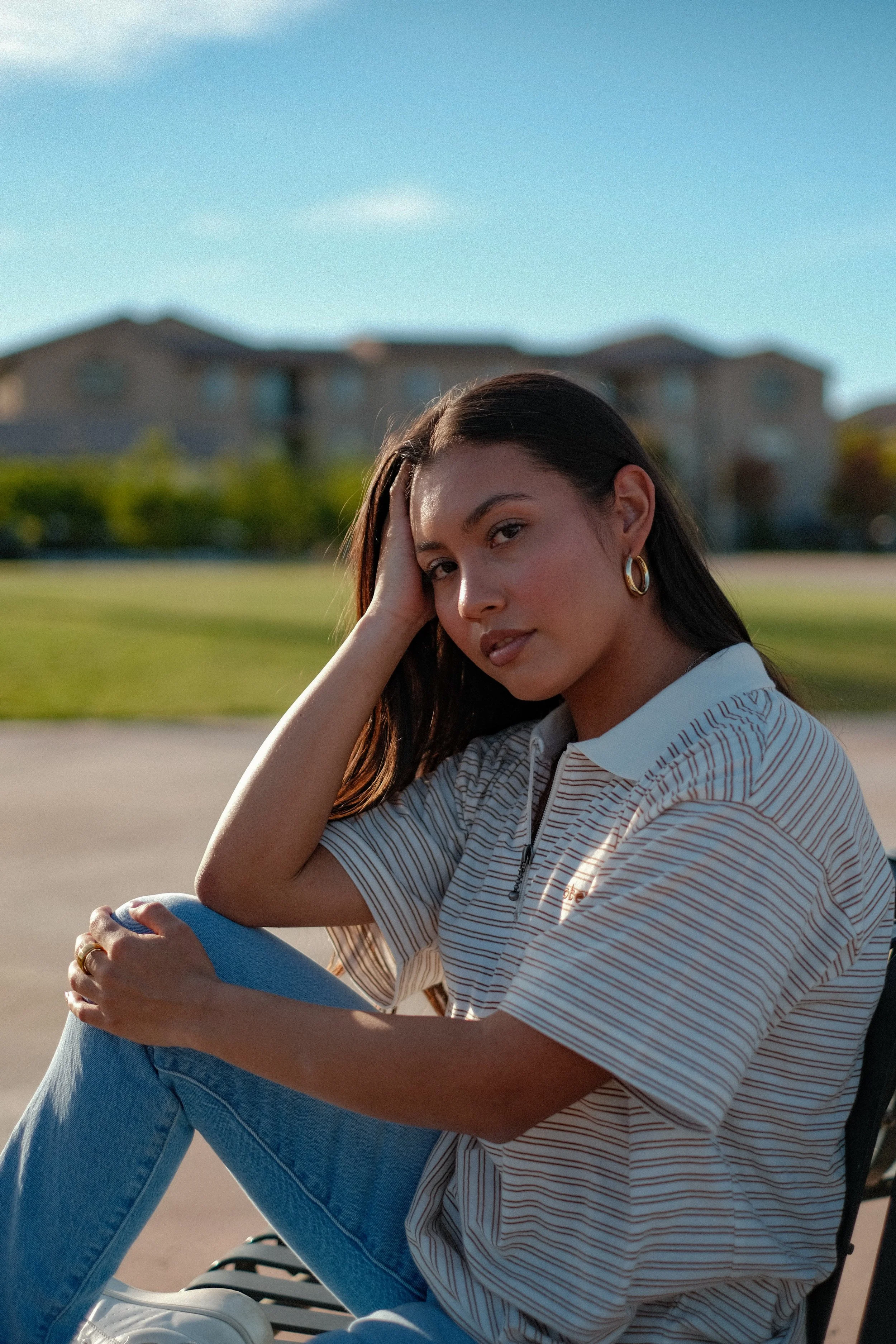 A young woman with long dark hair wearing a striped short-sleeve polo shirt and blue jeans, sitting outdoors on a bench during daytime, with her right hand resting on her head. She is looking into the camera with a neutral expression, and the backgro