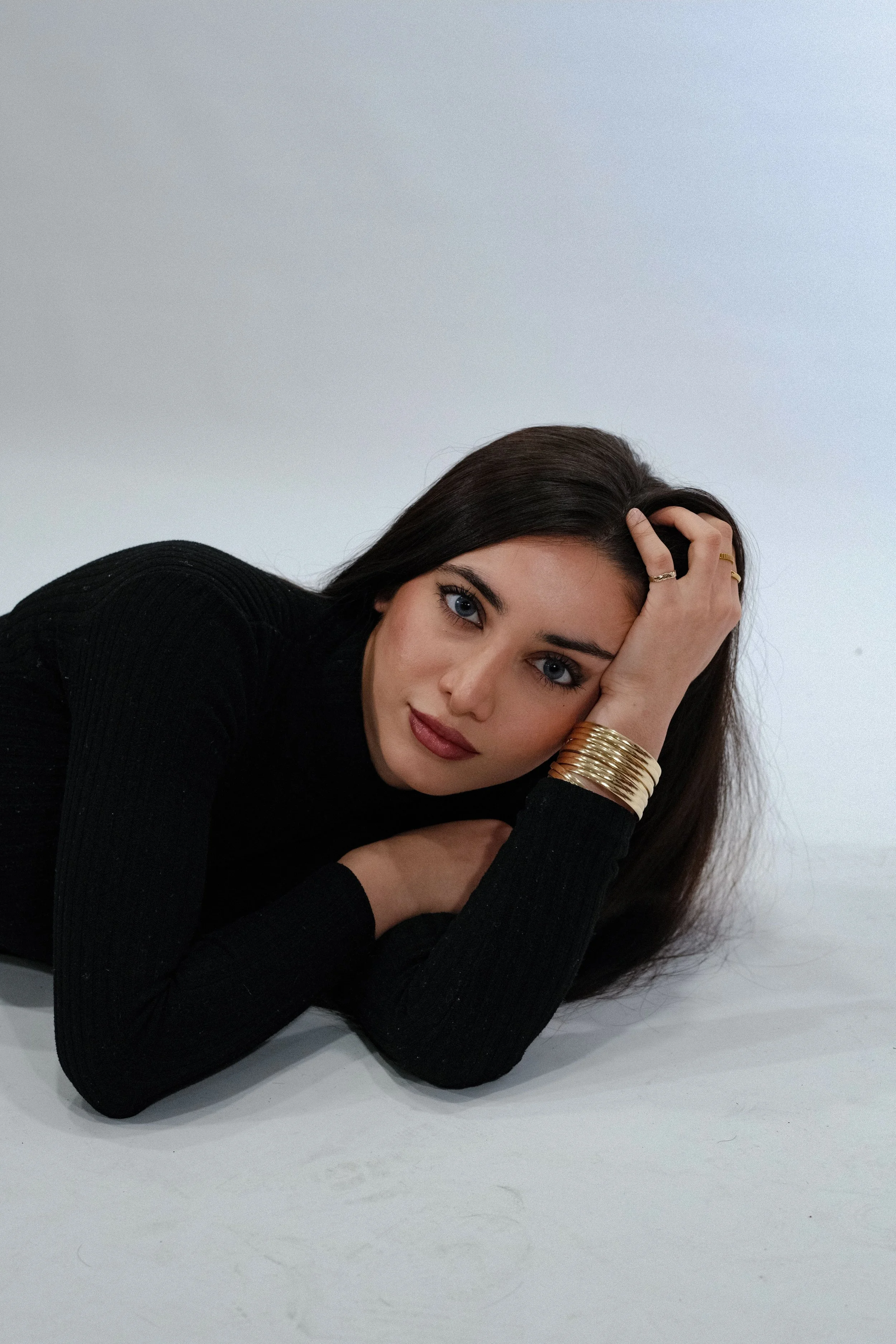 A woman with dark hair and blue eyes lying on her side on a white surface, wearing a black long-sleeve top and gold jewelry, looking at the camera.