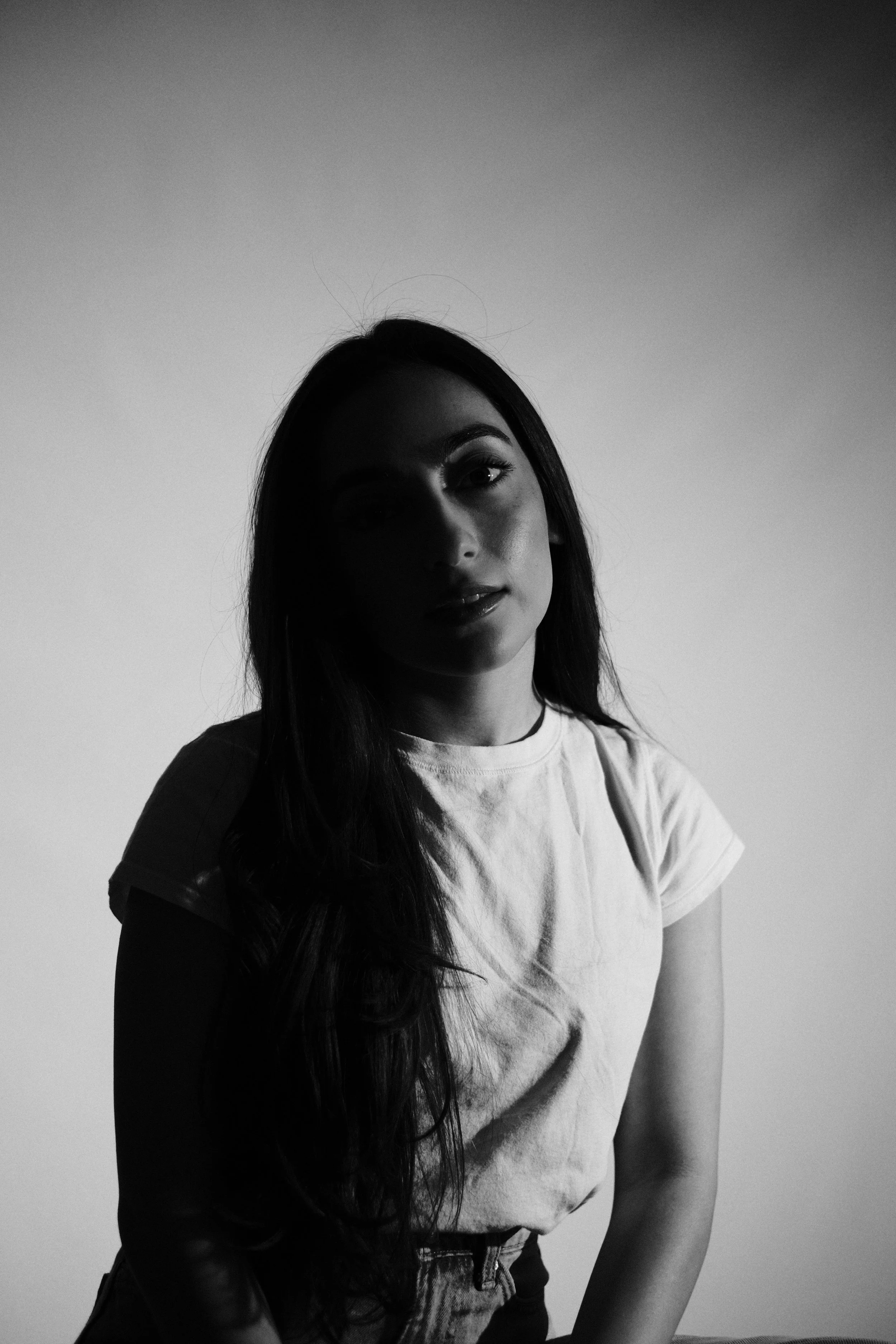 Black and white photo of a woman with long dark hair, wearing a white t-shirt, looking into the camera with a half-smile.