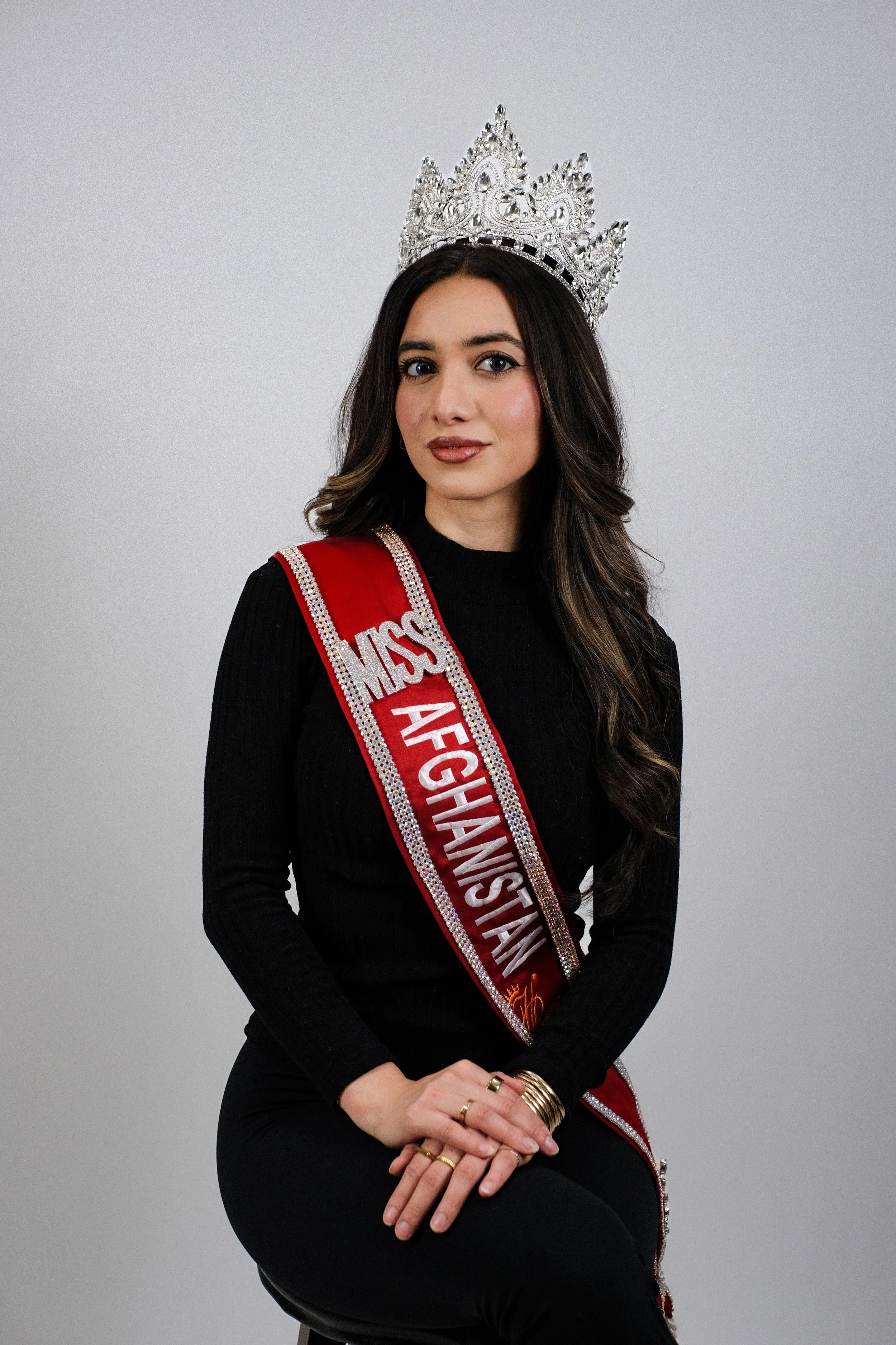 Miss Afghanistan wearing a crown, a black long-sleeve top, black pants, and a sash with 'MISS AFGHANISTAN' written on it, sitting on a stool against a plain background.