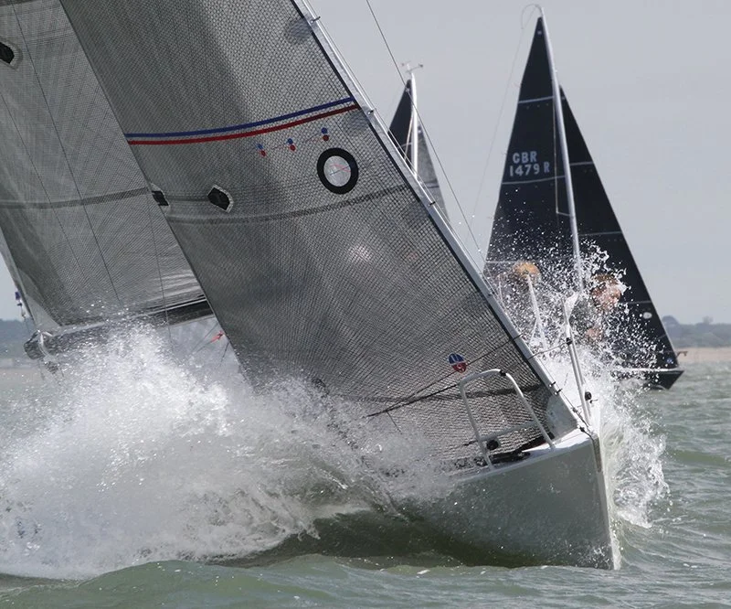 Two sailboats racing on the water, leaning into the turn with water splashing against their hulls.