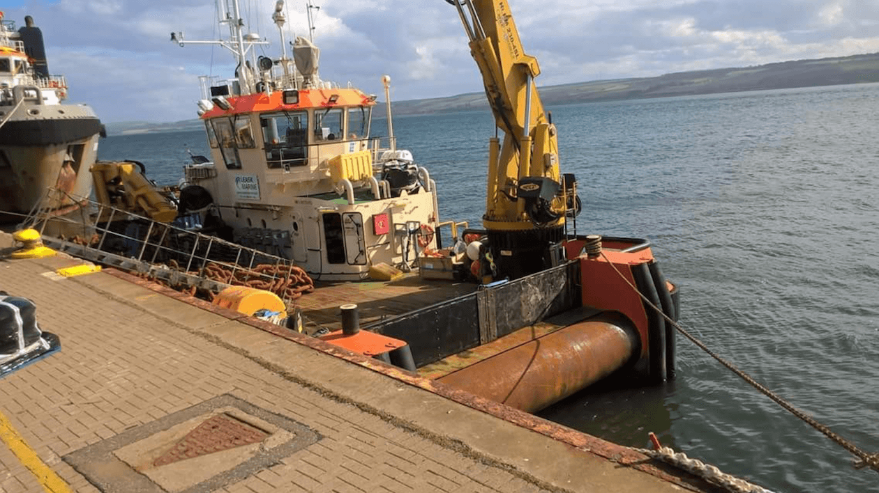 A boat docked at a pier with a crane on board, rusted buoys, all surrounded by water and hills in the background.