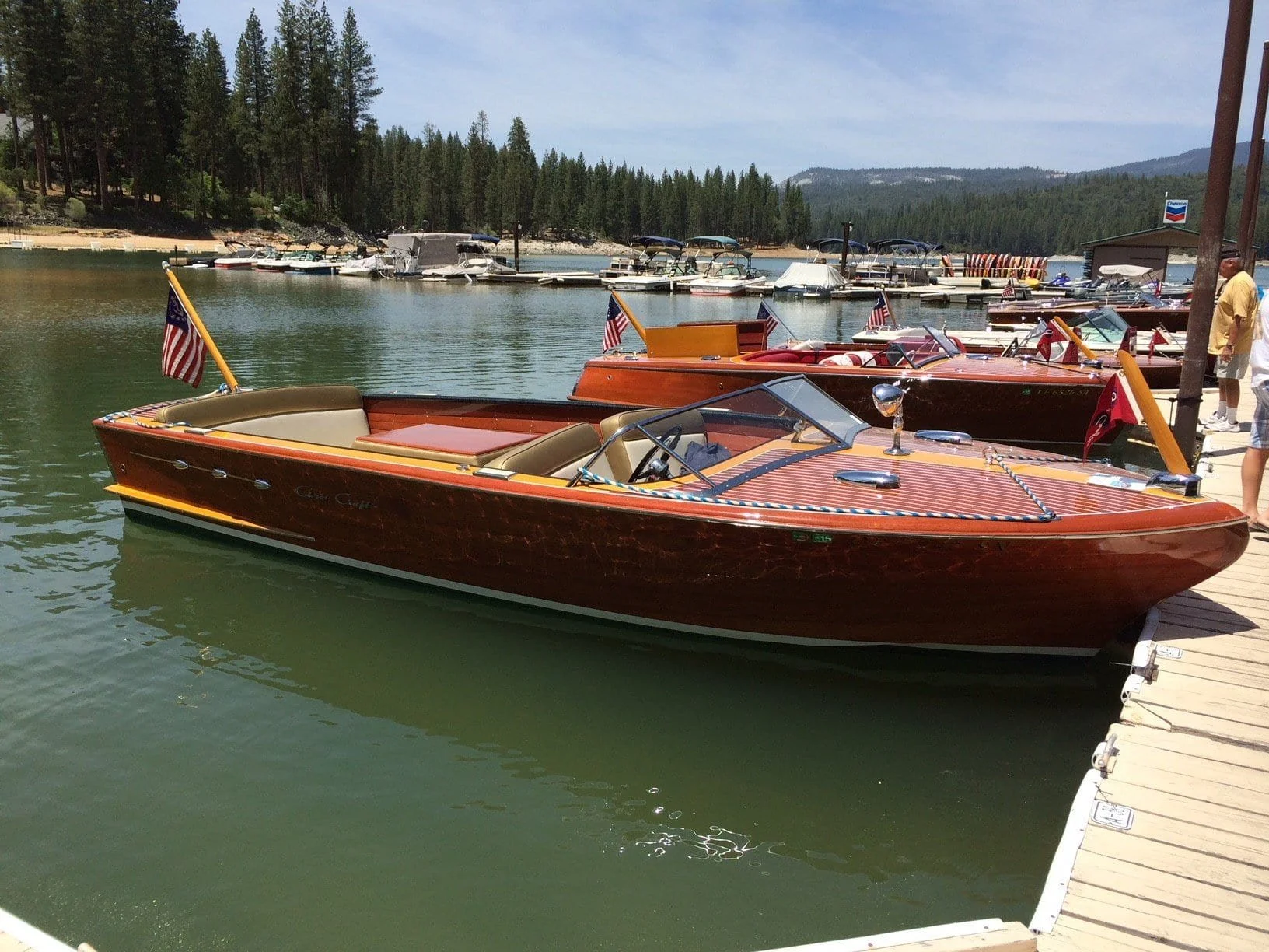 A wooden speedboat docked at a marina with other boats in the background, surrounded by trees and mountains.