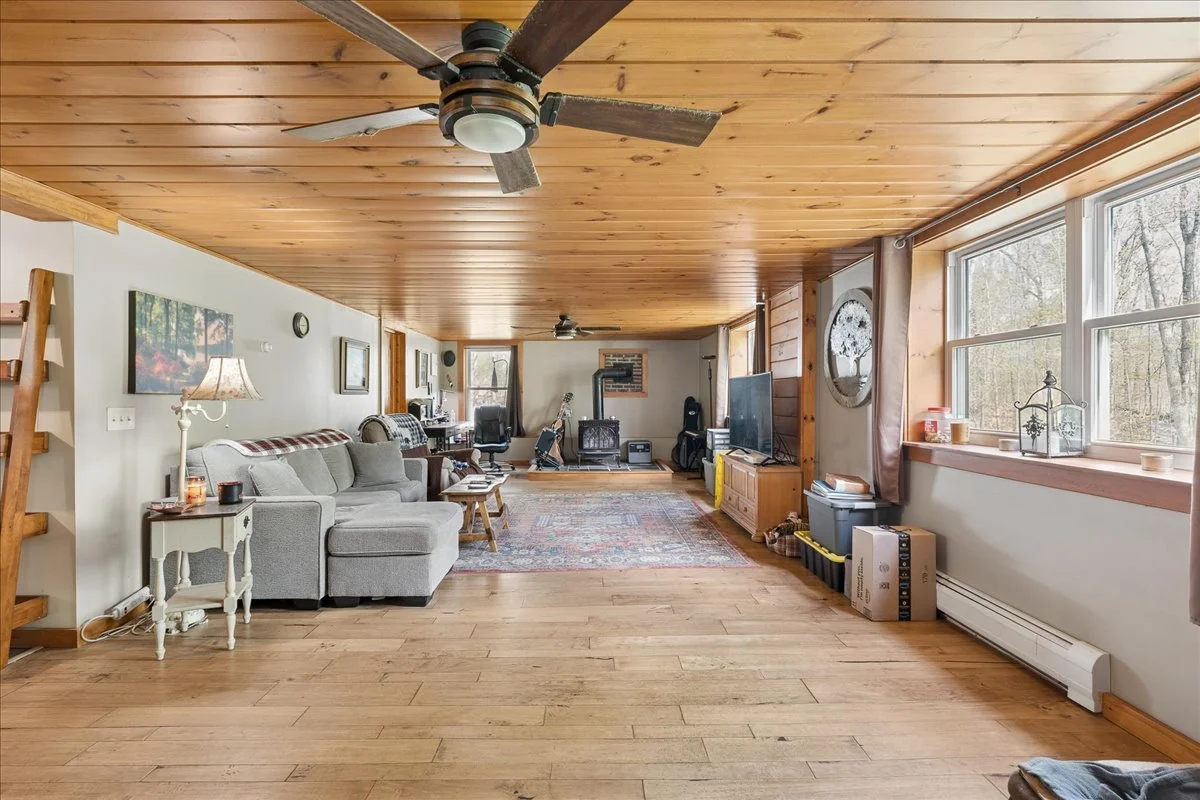 Spacious living room with wood-paneled ceiling and hardwood floors, furnished with a gray sectional sofa, side tables, a large flat-screen TV, and a wood stove. Large windows let in natural light, and various decorations are visible throughout the room.