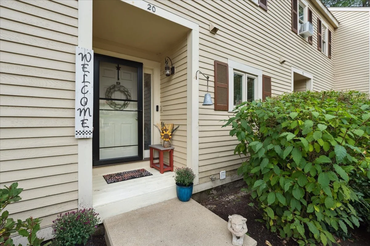 Front porch of a beige house with a welcome sign, small potted plant, sunflower arrangement, and a decorative lion's head sculpture outside. A bush and some flowers are nearby.