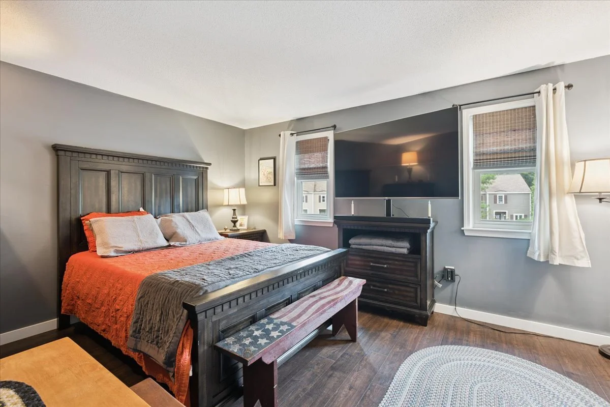 Bedroom with a dark wooden bed, orange bedspread, gray pillows, a nightstand with a lamp, and twin windows with white curtains. A large flat-screen TV is mounted above a dark wooden dresser.