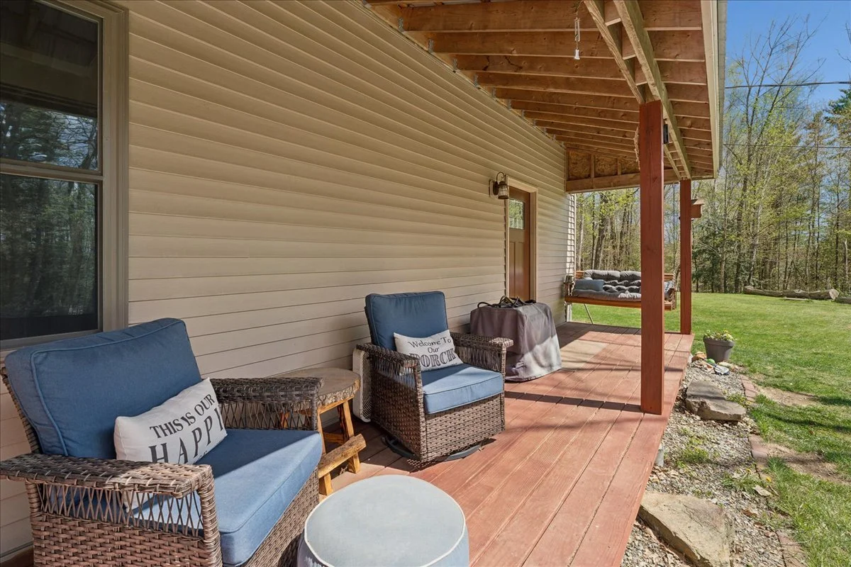 A covered porch with wicker chairs that have blue cushions and pillows, one with a sign that reads 'This is our happy place.' There are some small tables and a covered grill or smoker. The porch overlooks a grassy yard with trees in the background.