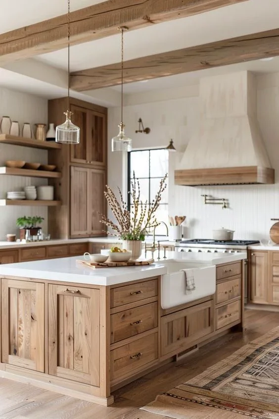 A rustic kitchen with wooden cabinets and a large white island counter. Open shelving displays dishes and plants, with a window and stove in the background. Decor includes a vase with dried flowers.