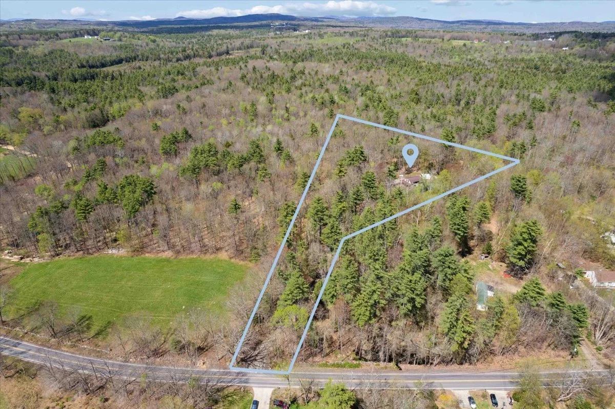 Aerial view of a wooded property with a house and driveway, outlined with a blue border, in a rural area with neighboring houses, a road in the foreground, and open fields in the distance.