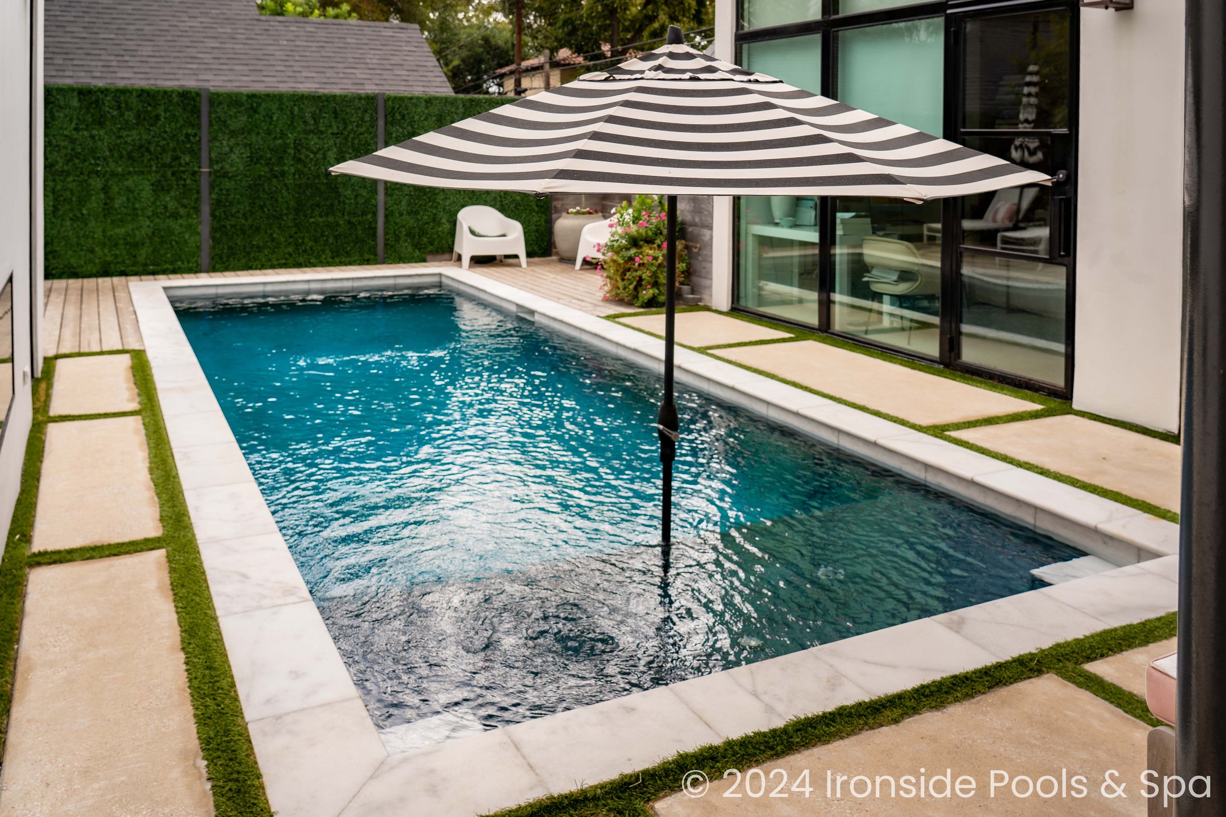 Modern backyard swimming pool with striped umbrella, deck chairs, and privacy hedge. Contemporary design with glass windows and greenery. © 2024 Ironside Pools & Spa
