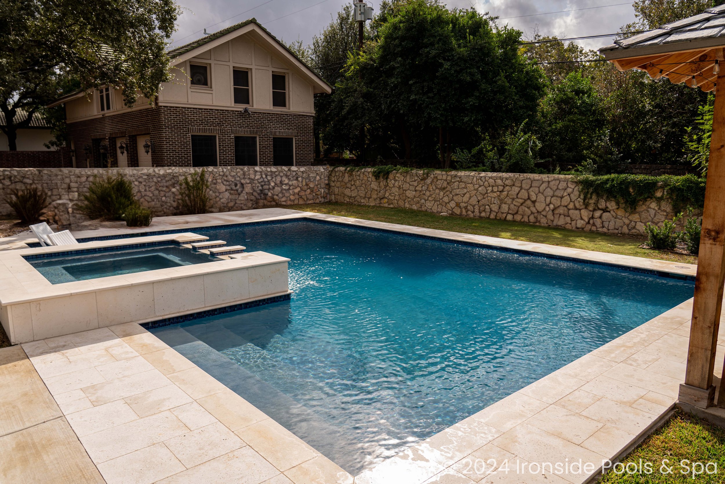 Backyard with a rectangular swimming pool, adjacent hot tub, stone deck, and a backdrop of a two-story house and stone wall.