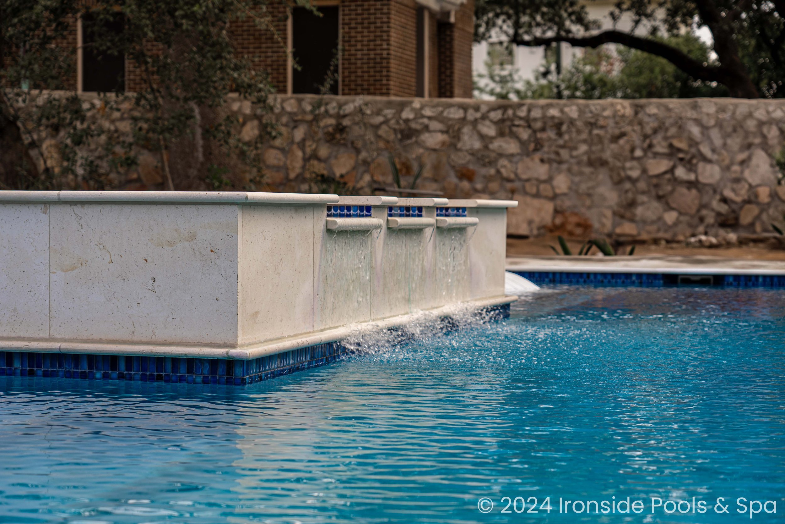 Swimming pool with cascading water feature and stone wall in the background.