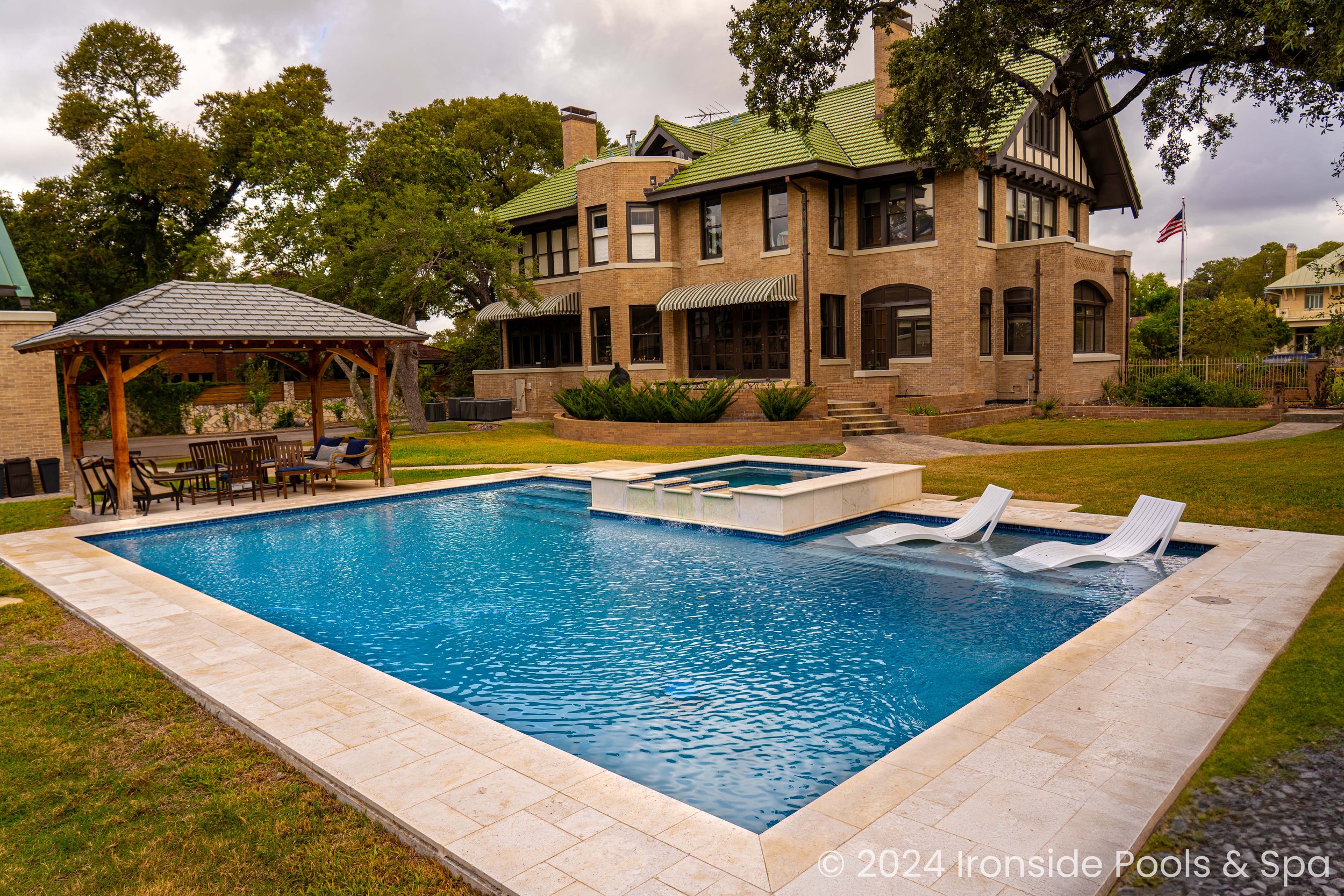Elegant backyard with a large pool, hot tub, and sun loungers by a mansion. There's a wooden seating area with a table under a gazebo on the lawn.