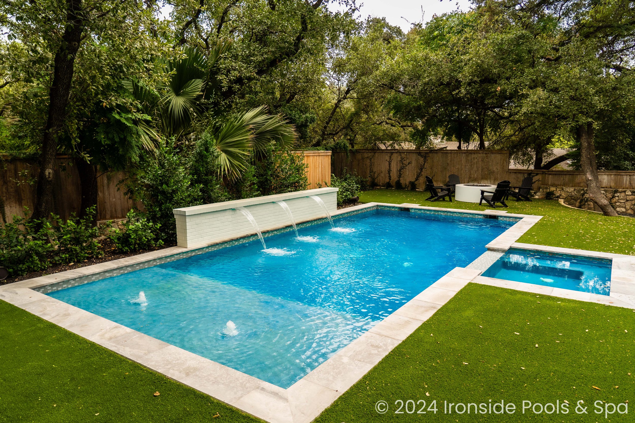 Backyard with a rectangular swimming pool featuring three water fountains, surrounded by green artificial turf. There's a hot tub adjacent to the pool and a small seating area with black chairs and a white table, set near trees and a wooden fence.