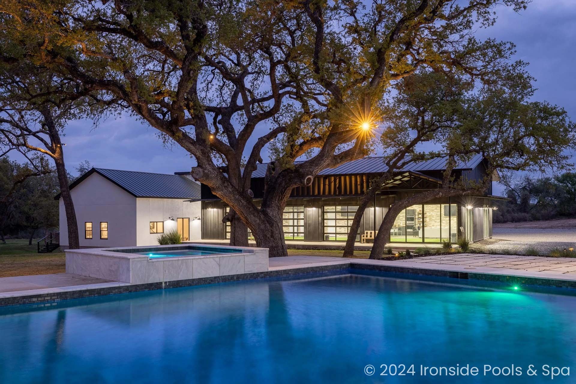 Modern house with large windows and outdoor pool, surrounded by trees at dusk.