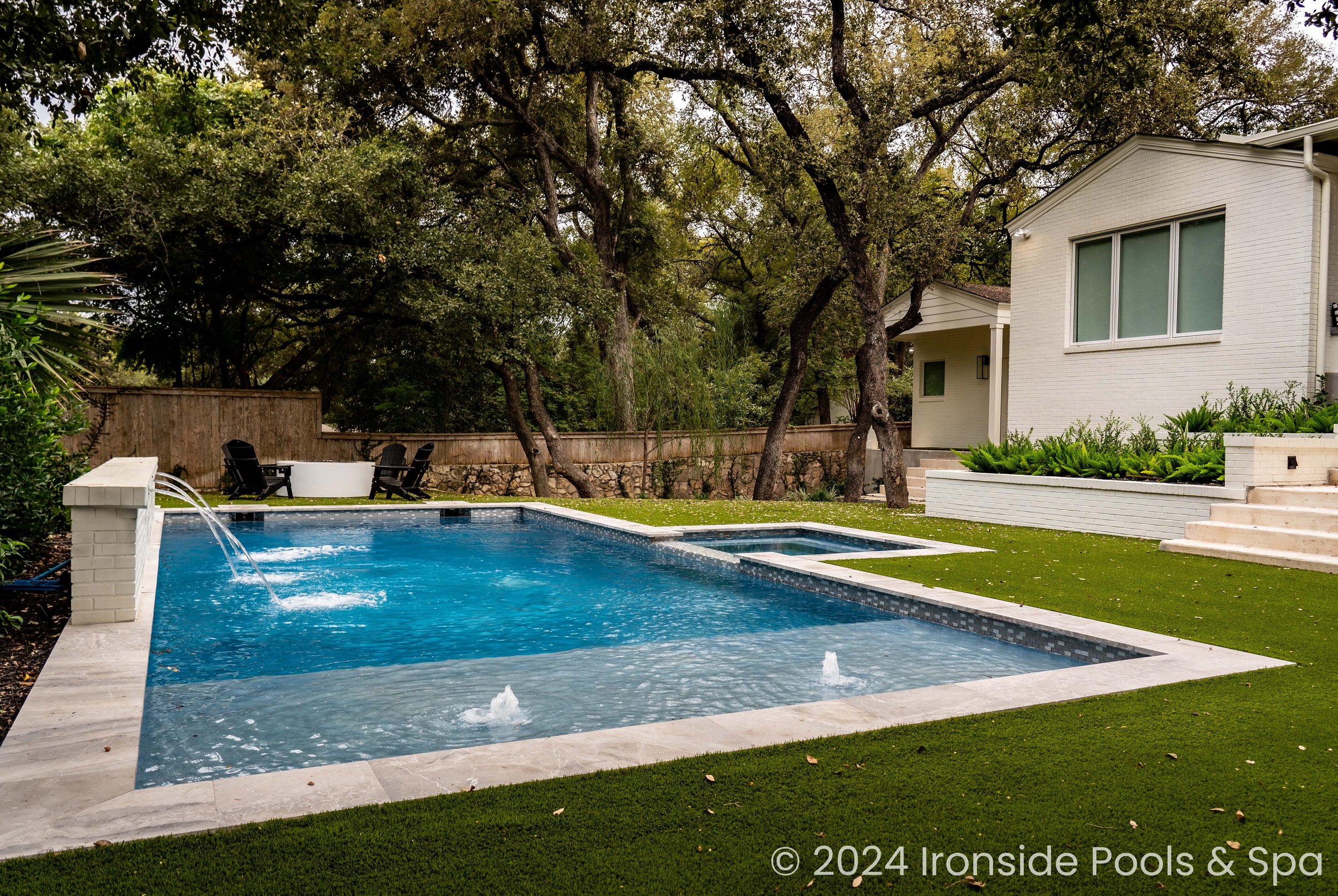 Backyard with a modern swimming pool, a small hot tub, and water features, surrounded by artificial grass and trees, adjacent to a white house.