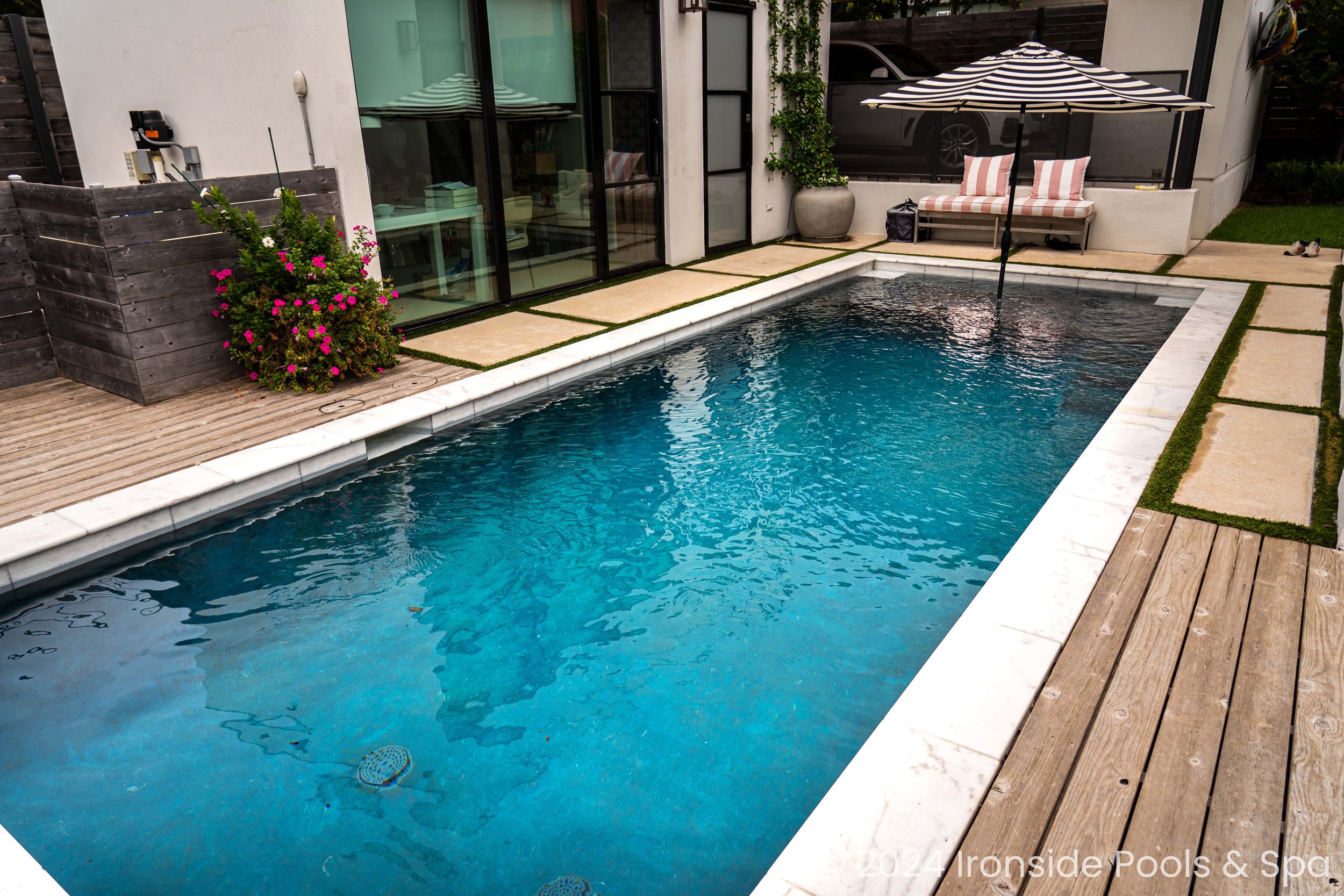 Modern backyard with a rectangular swimming pool, surrounded by a wooden deck and stone slabs, with a bench and a striped umbrella in the background.