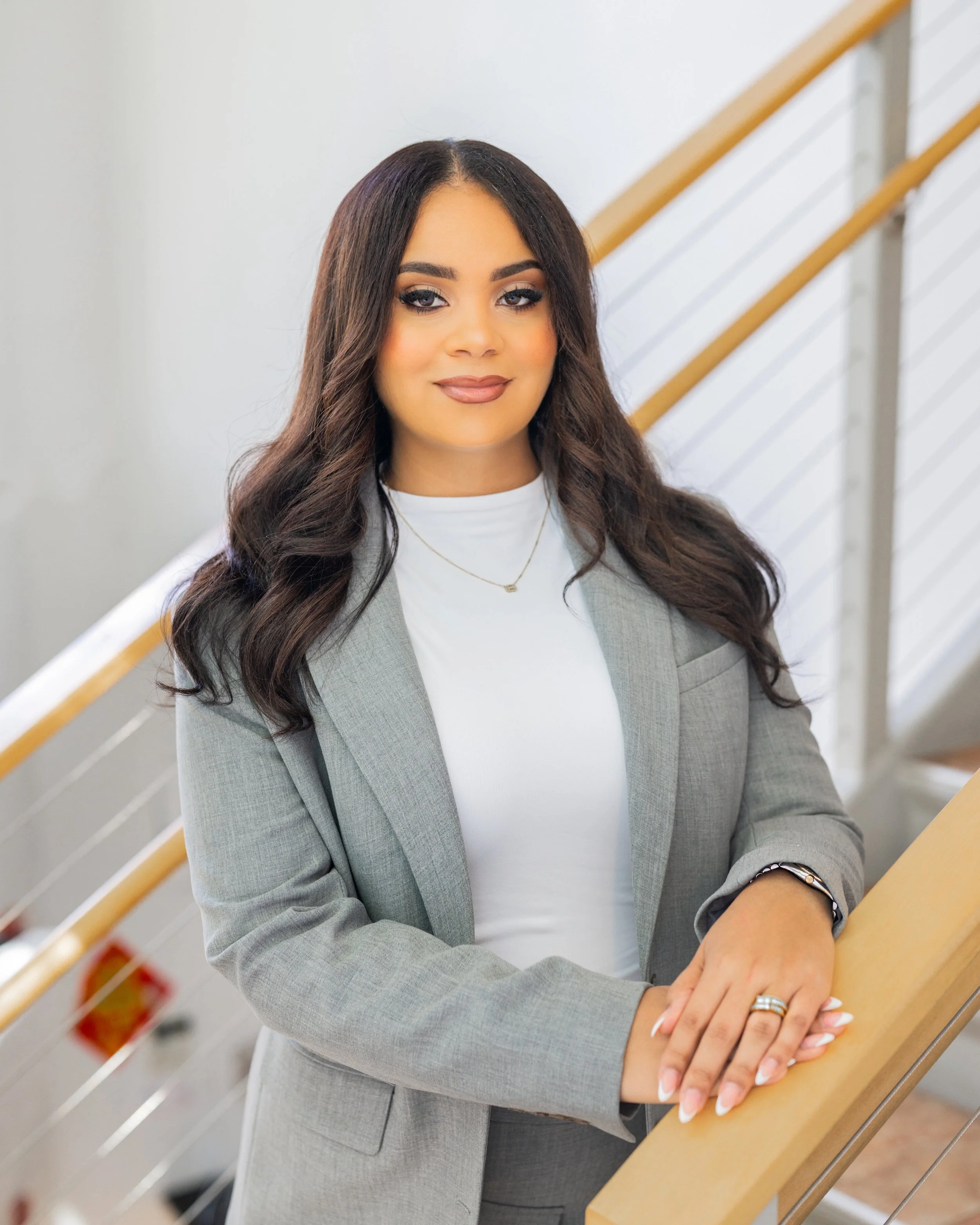 A clean, bright photo of a 30 year old woman lawyer standing at the bottom of a staircase. She has hands folded over the banister. She is wearing a high neck shirt with a blazer over top. Her hair is mid length.  She has a smirk showing no teeth.