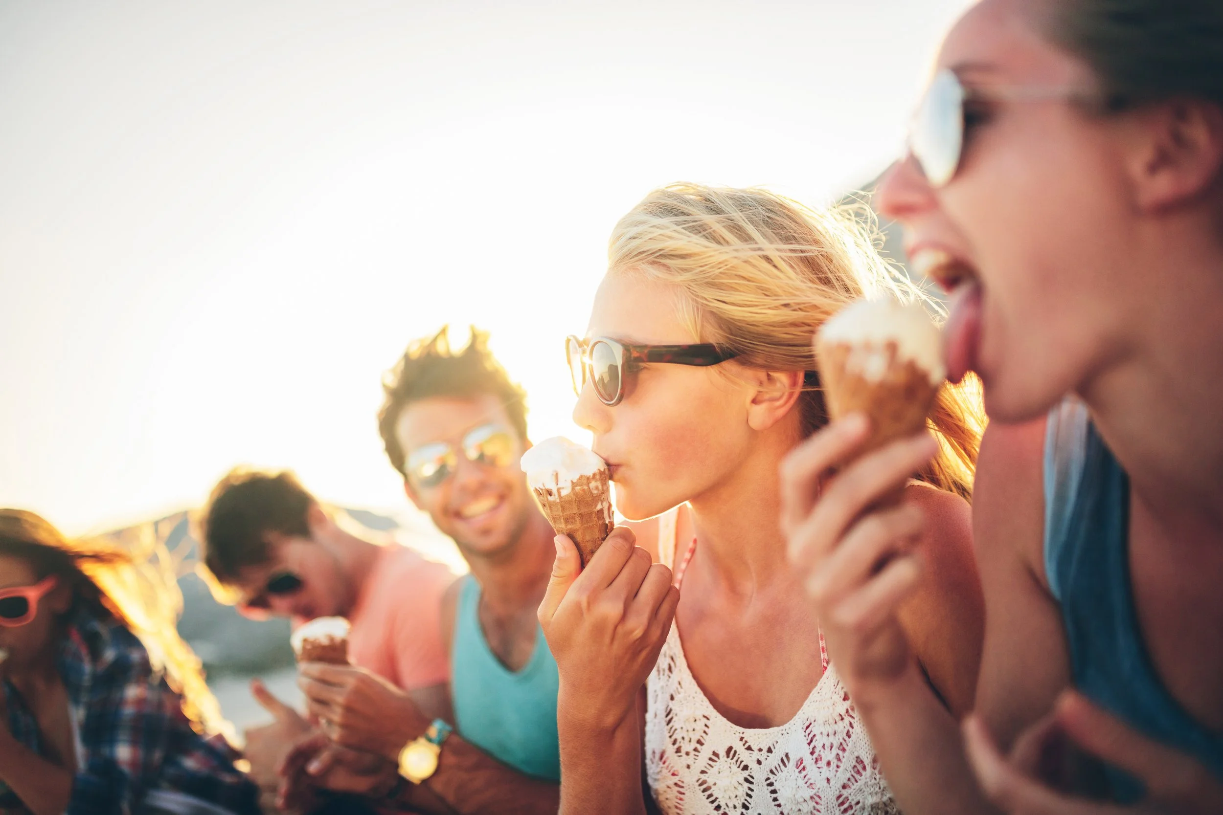 Group of friends enjoying ice cream cones outdoors at sunset, wearing sunglasses and casual clothing.