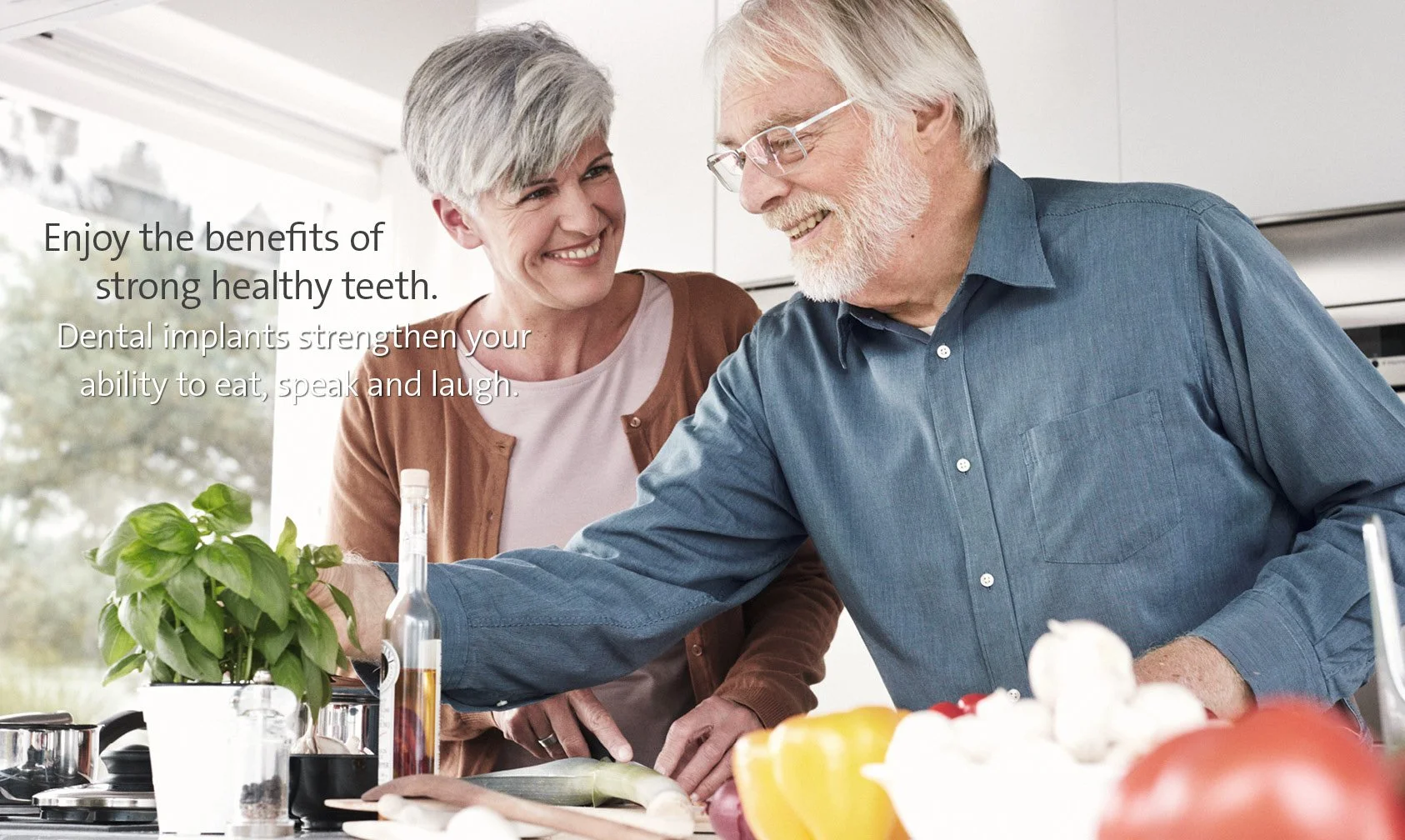 An elderly man and woman smiling in a kitchen, with the man reaching toward a potted herb, surrounded by vegetables and cooking ingredients.
