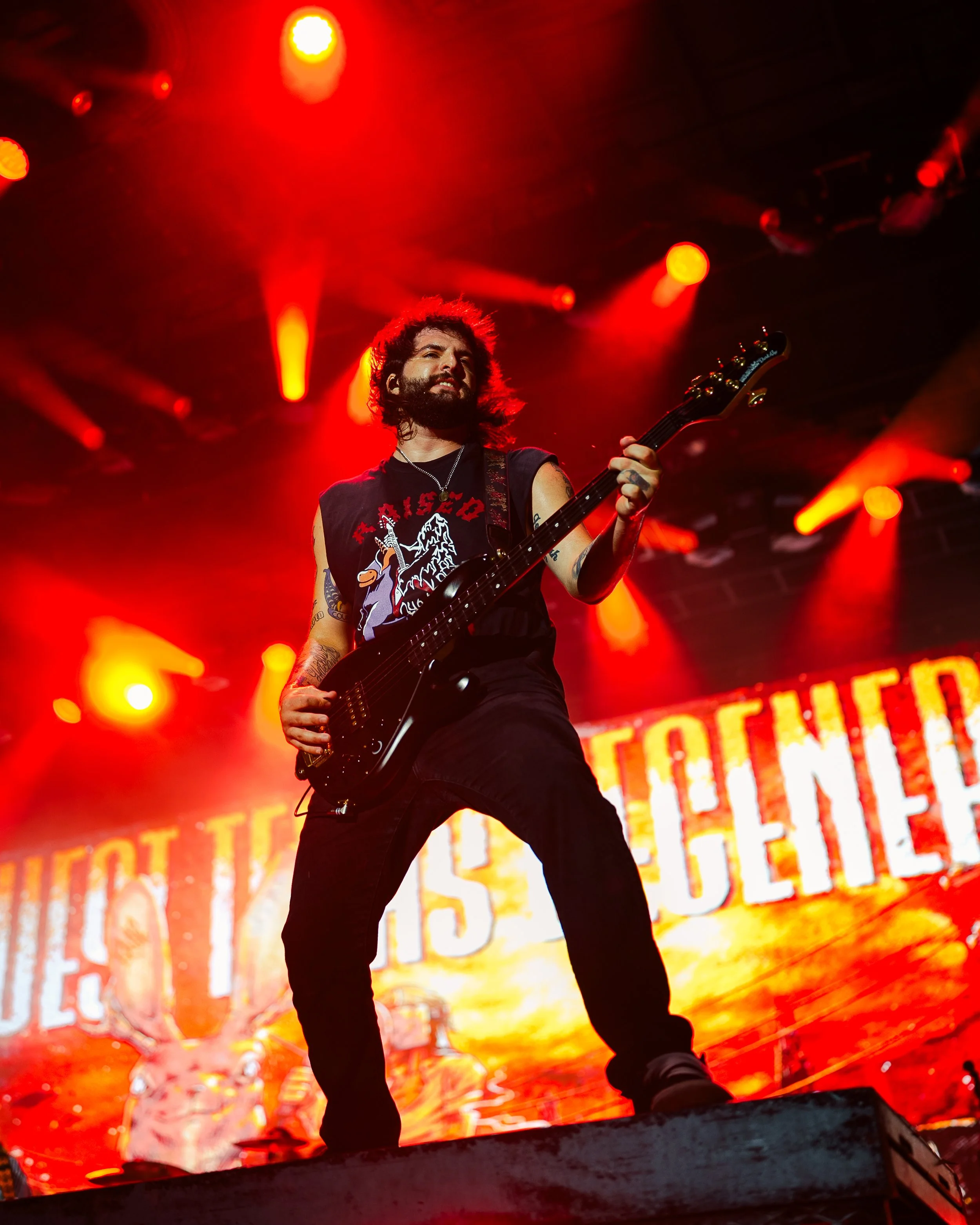 A musician playing guitar on stage with red and orange stage lights and a blurry background of a digital screen displaying text.
