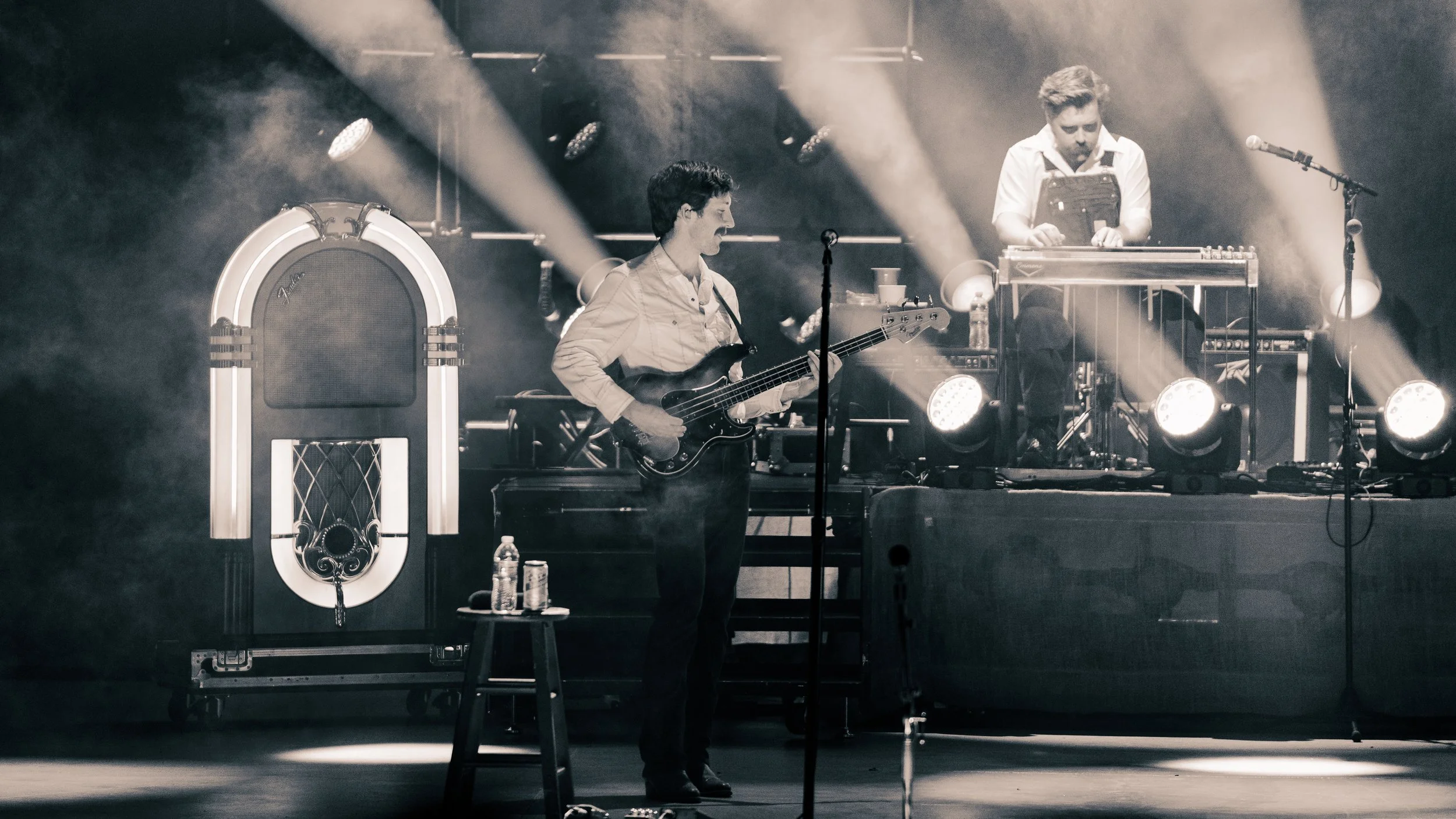 A black-and-white photo of two musicians on stage, one playing a bass guitar and the other operating electronic equipment, with stage lights shining behind them.