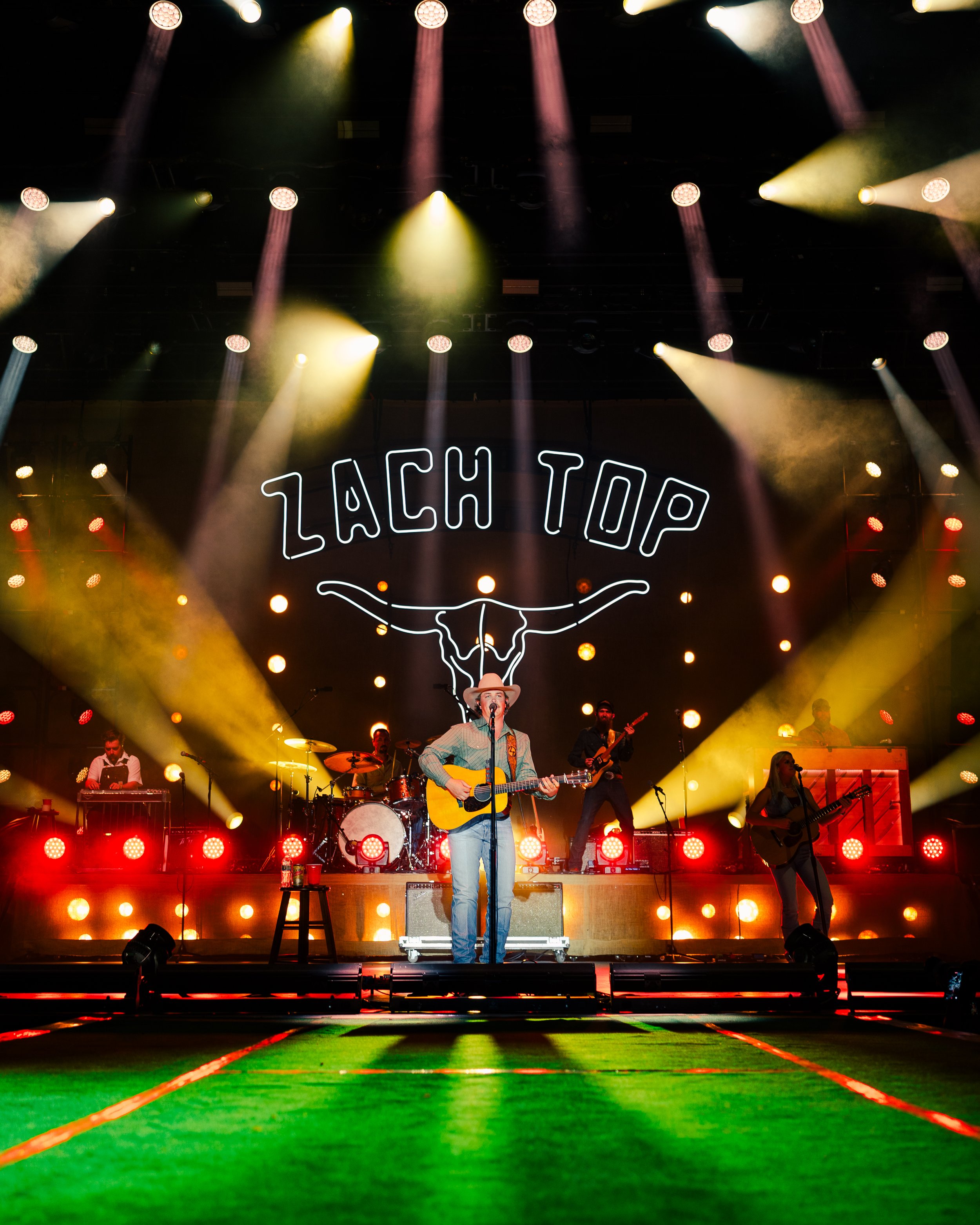 Performer with cowboy hat singing on stage with a band and neon 'LACH TOP' sign behind him, colorful stage lights, and a green floor in the foreground.